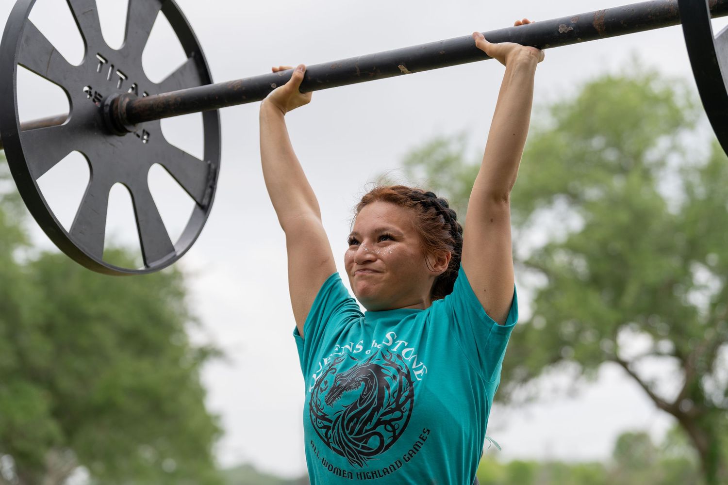 Woman in teal shirt lifting barbell overhead outdoors, showcasing strength training and weightlifting fitness.
