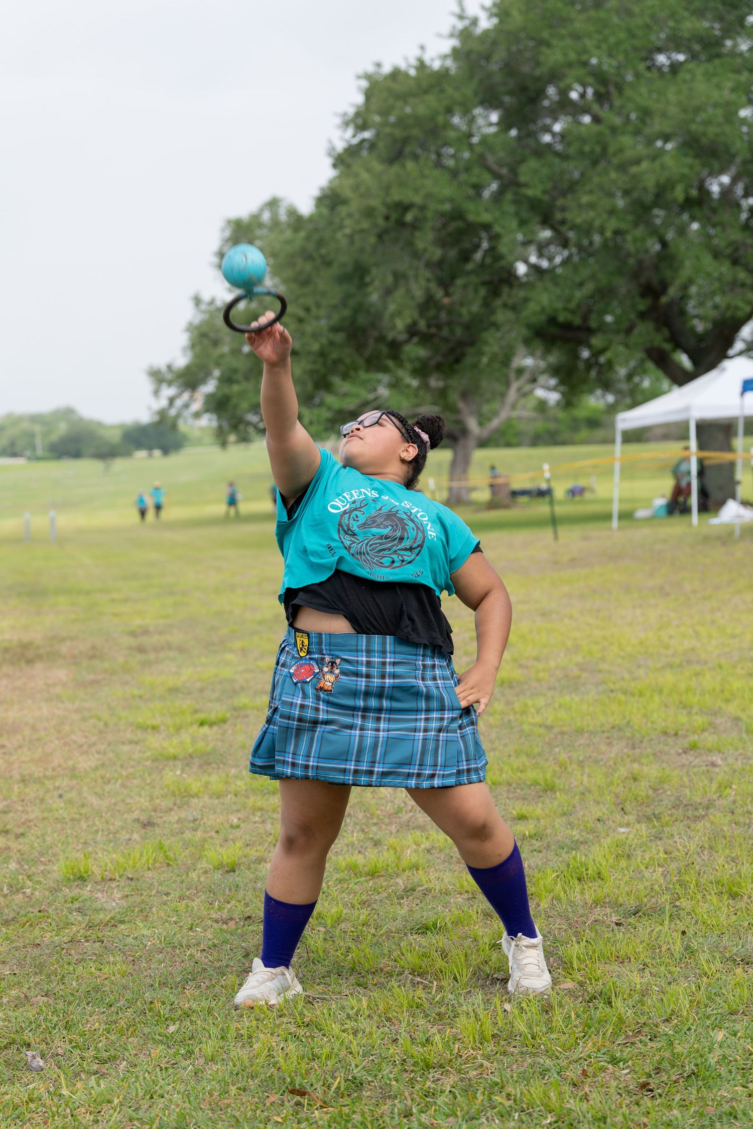 Woman in blue tartan skirt throws shot put at Highland Games outdoor competition on green field.