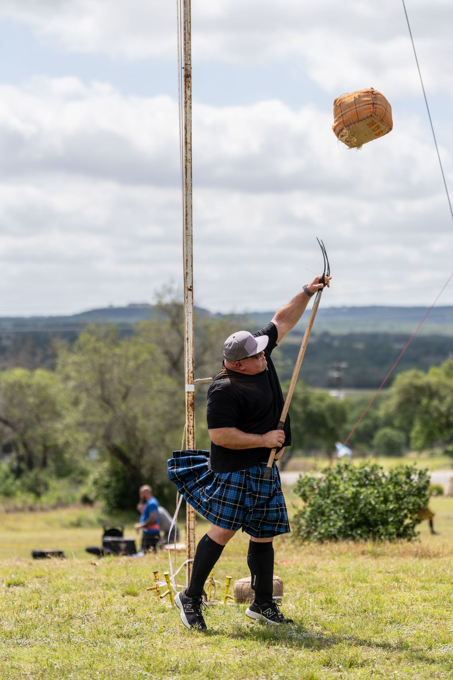 Man in blue tartan kilt competing in Highland Games sheaf toss event outdoors.