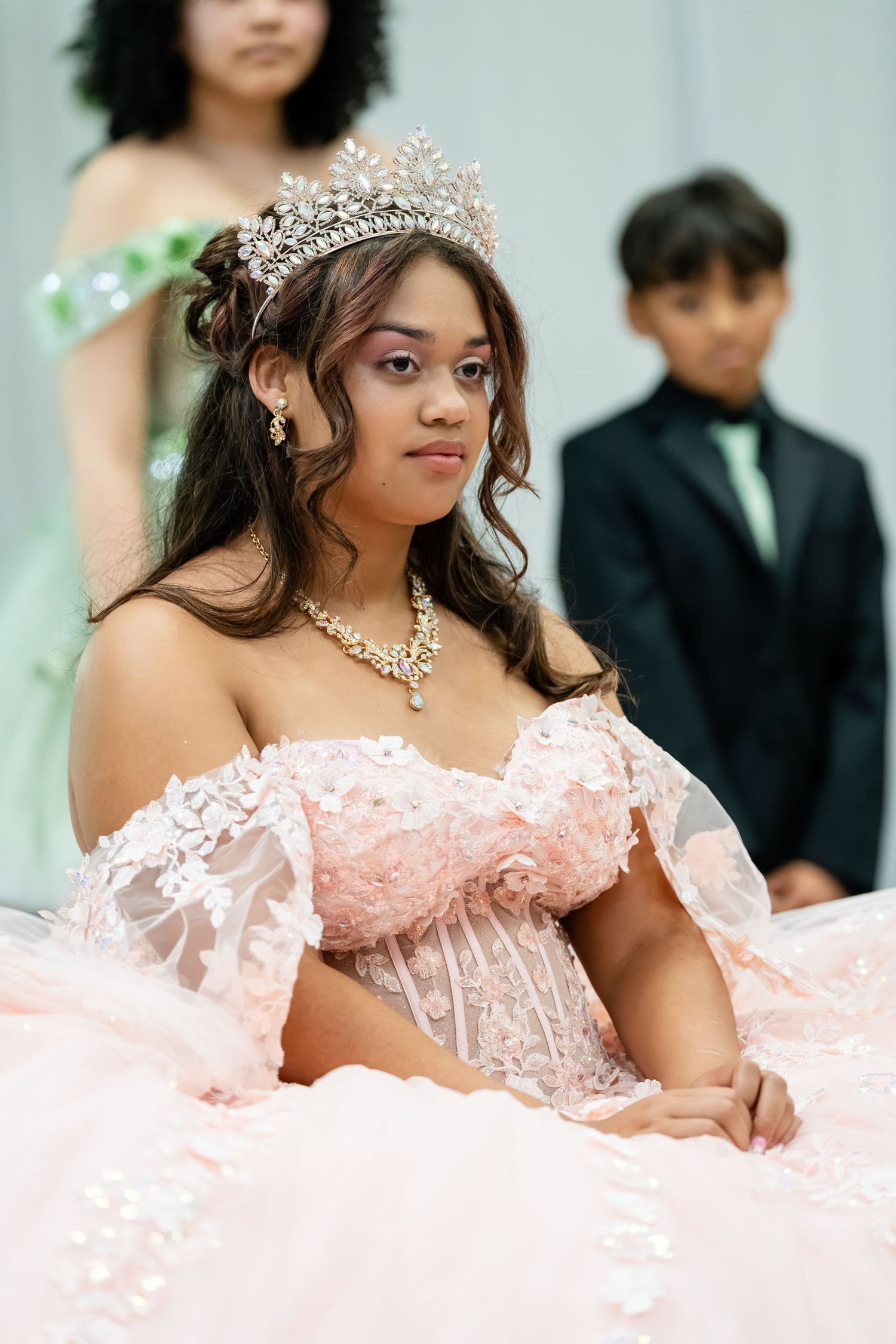 Young woman in pink off-shoulder quinceañera gown with crystal tiara and necklace poses elegantly at formal celebration.