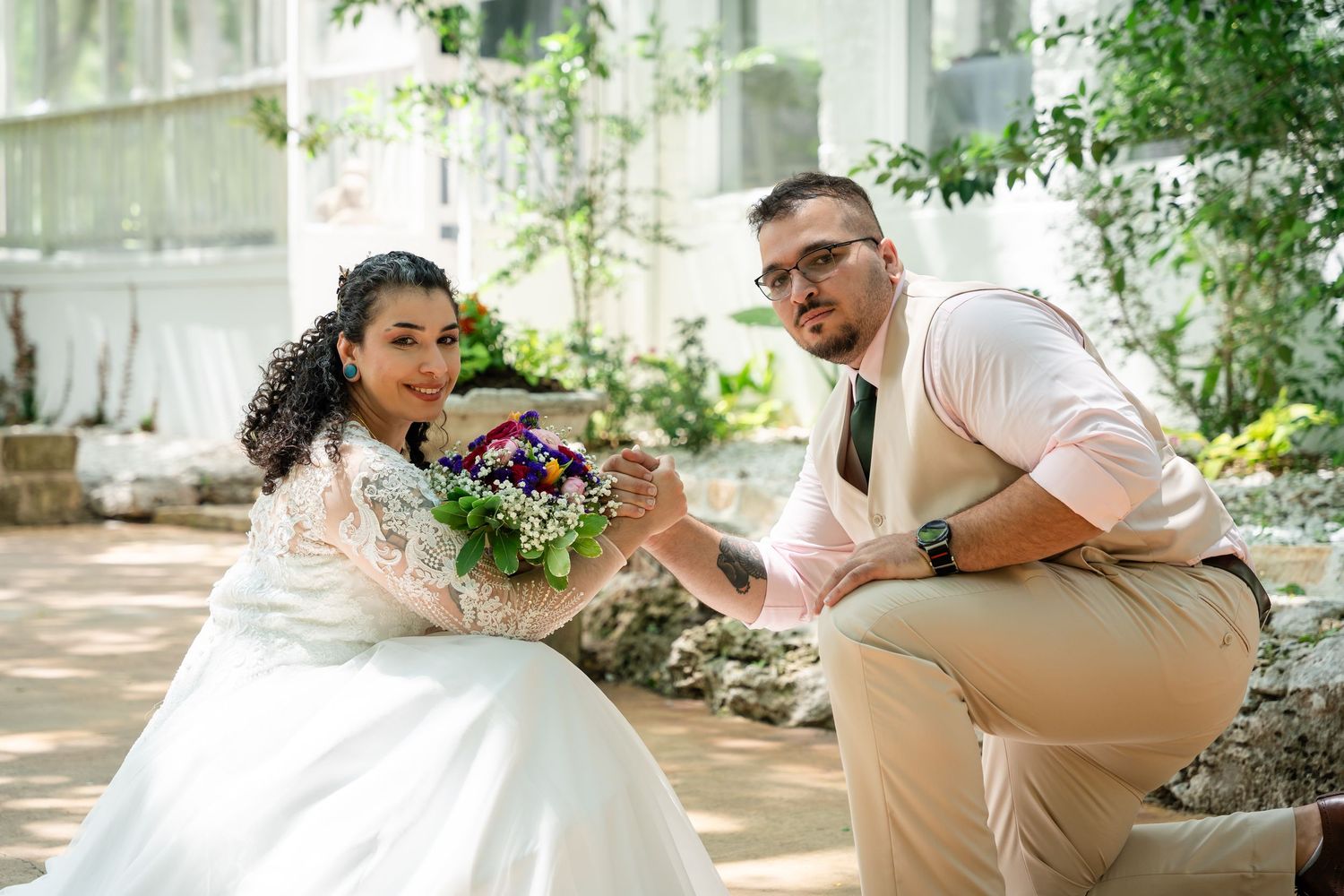 A bride and groom pose together in a greenhouse, she holds a colorful bouquet while he kneels beside her.