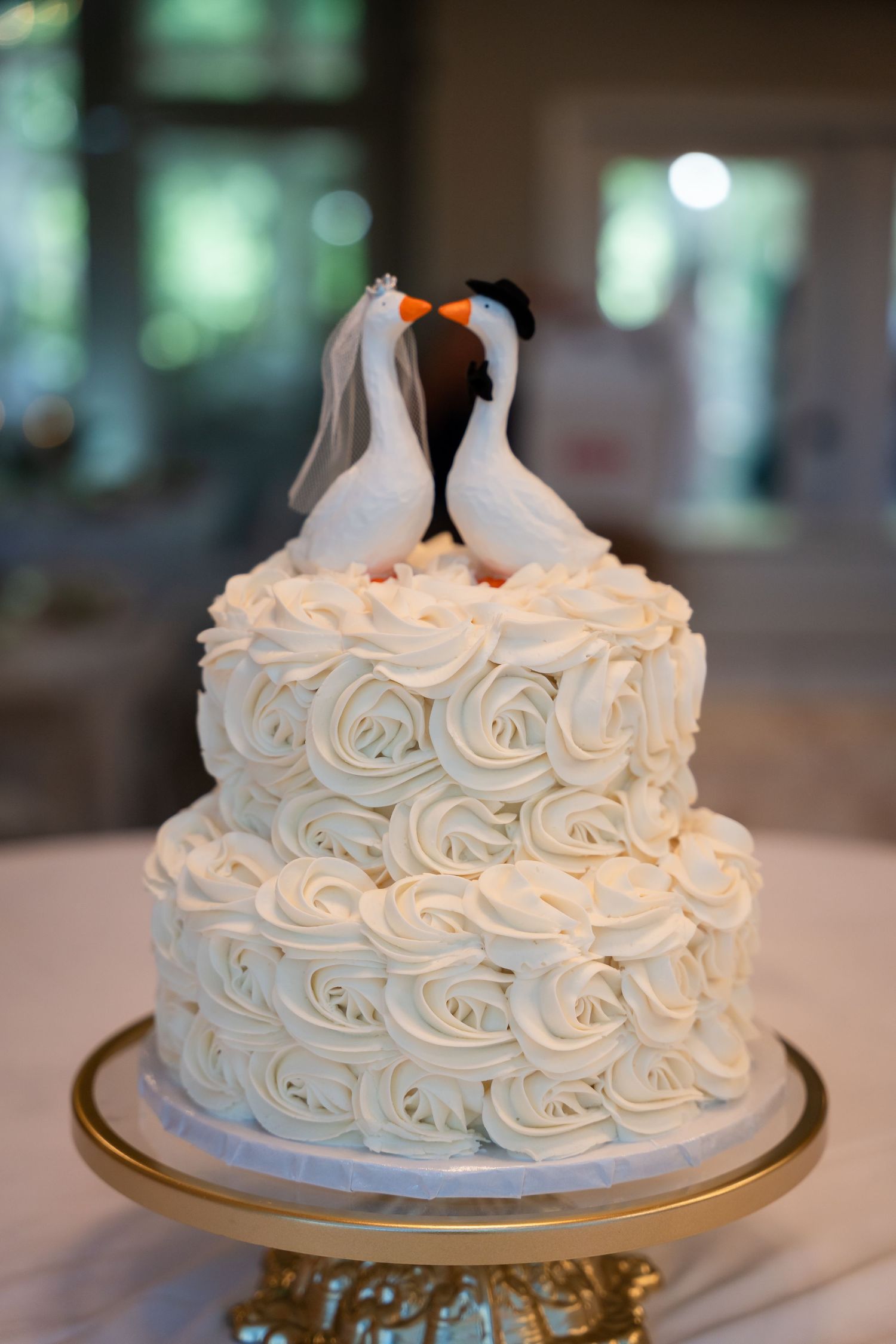 Two-tier white rosette wedding cake with penguin bride and groom cake toppers on a gold stand.