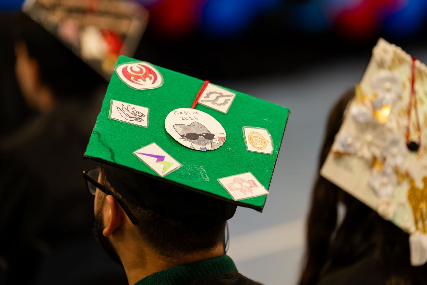 Green graduation cap decorated with colorful stickers and patches at a commencement ceremony.