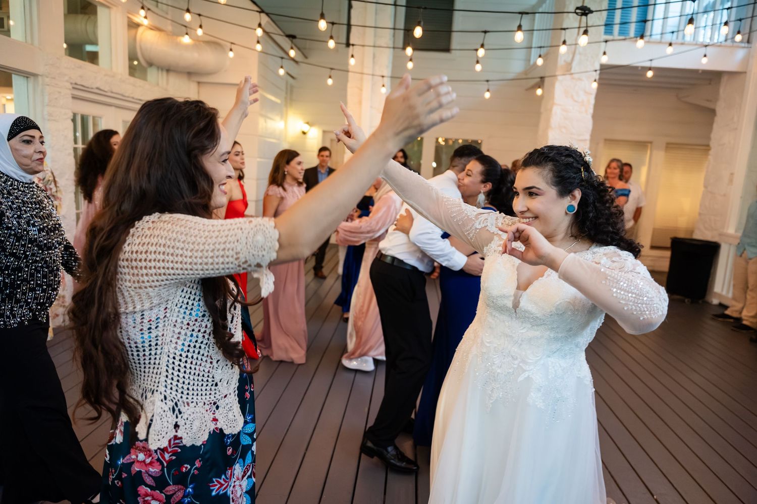 Bride in white gown dancing joyfully with guests at a wedding reception under string lights on a wooden dance floor.
