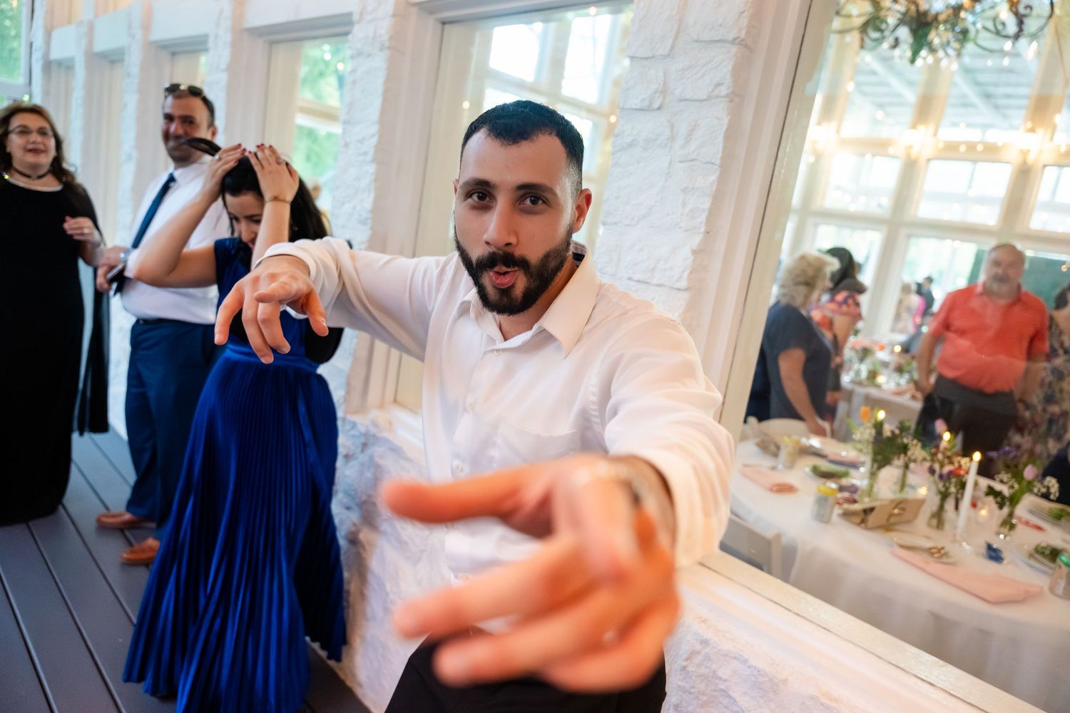 Man in white shirt dancing energetically at a wedding reception, reaching toward camera with guests and decorated tables behind him.