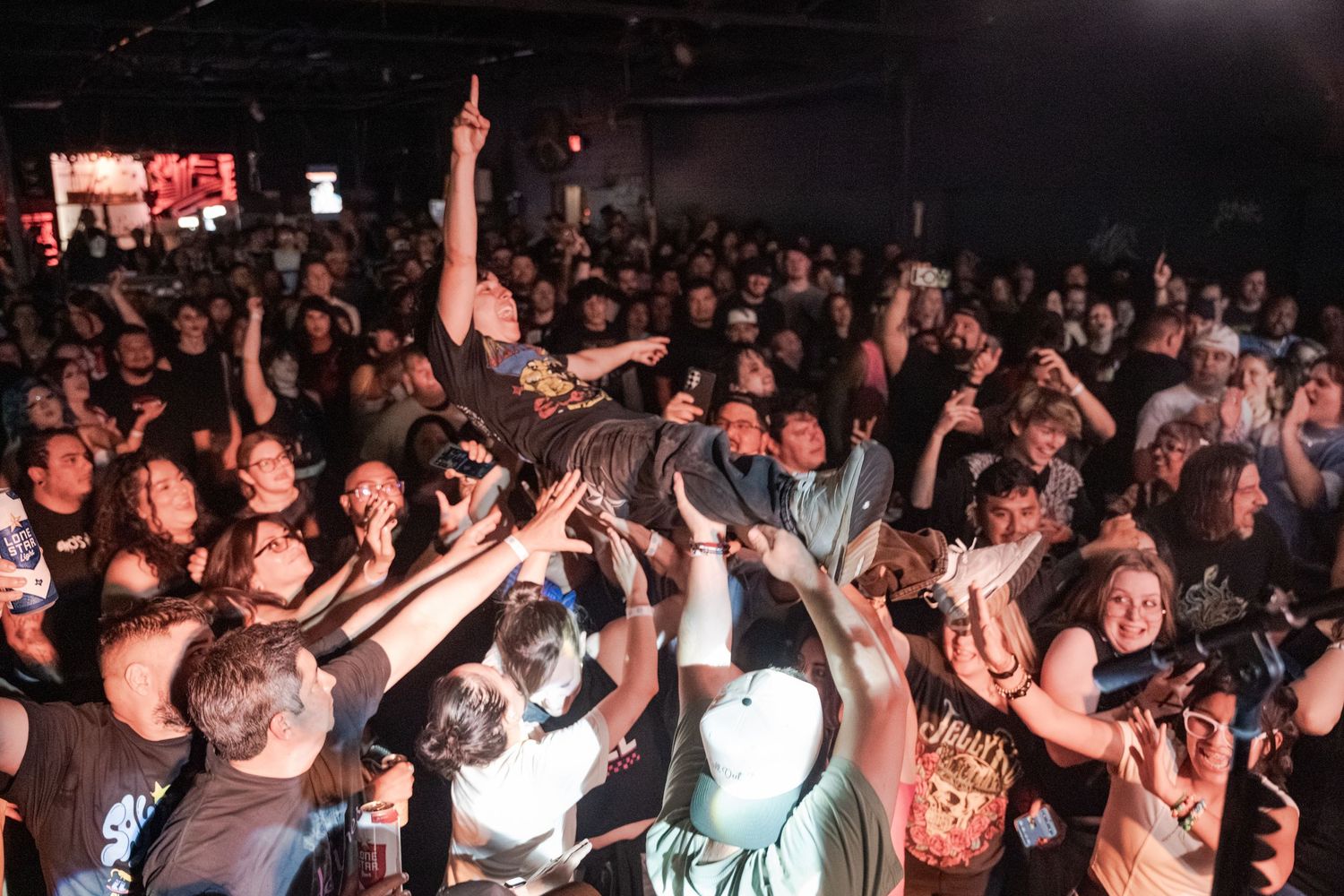 Energetic crowd at a packed rock concert venue with fans cheering and raising hands under bright stage lights.
