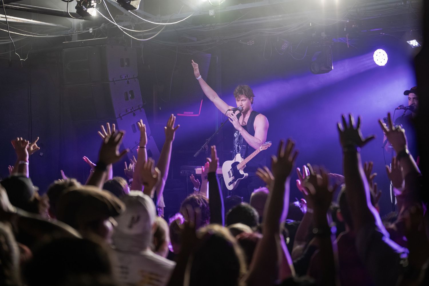 Guitarist performs on stage with one hand raised, energizing a crowd of fans with arms outstretched at a concert.