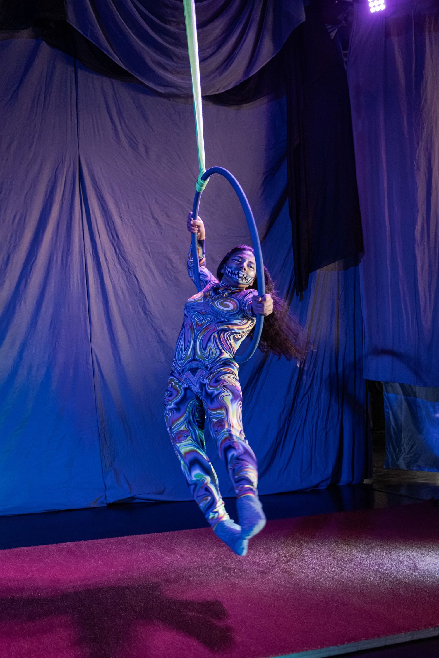 Aerial acrobat in colorful bodysuit performs on a hoop under blue stage lights against draped purple backdrop.