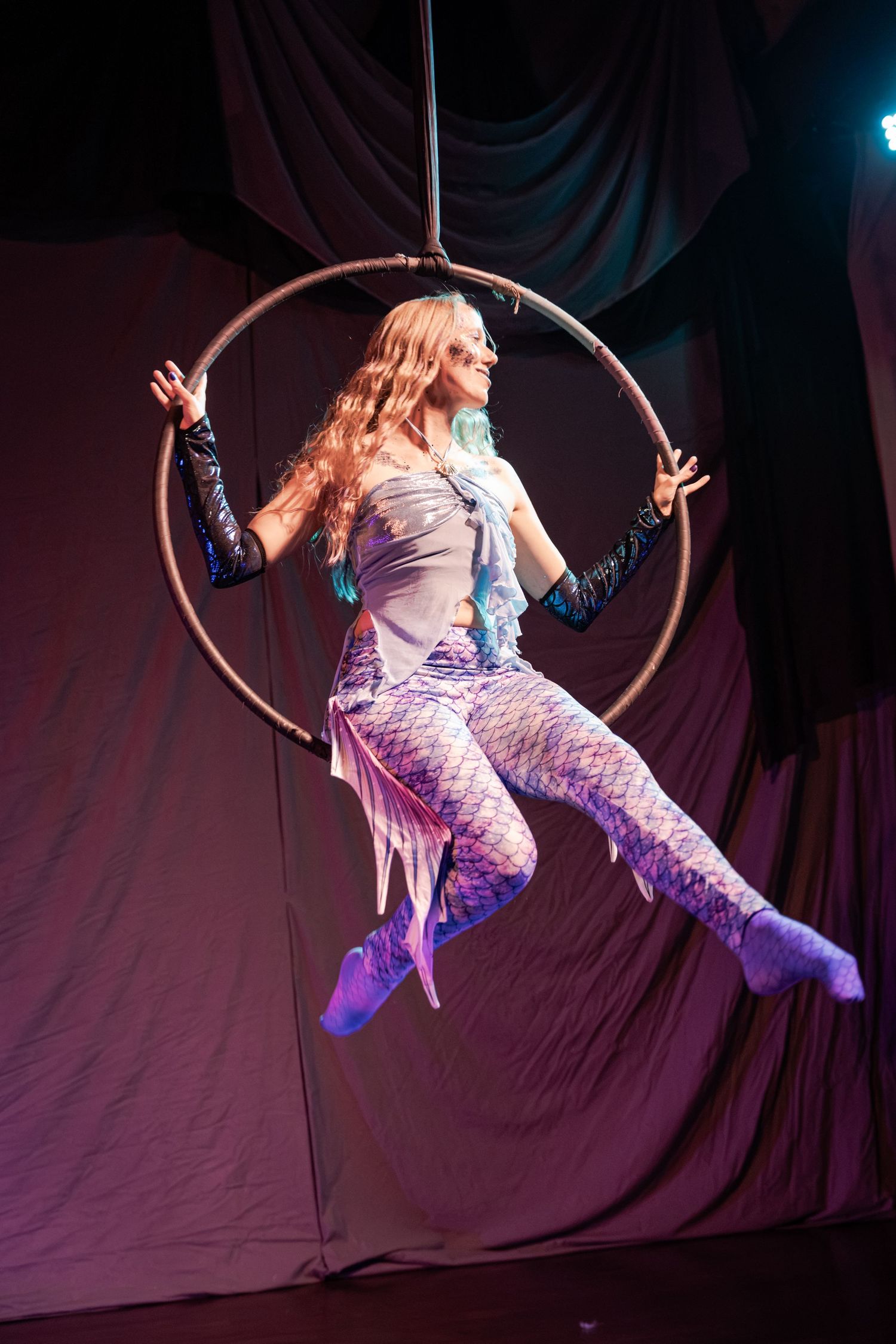 Aerial circus performer in mermaid costume poses gracefully inside a suspended hoop on a dramatic stage.