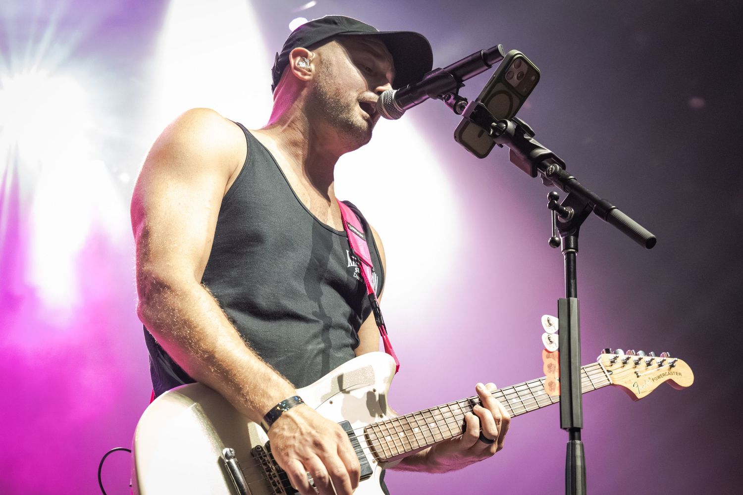 Male guitarist performing on stage with a white Fender guitar and microphone under purple stage lights.
