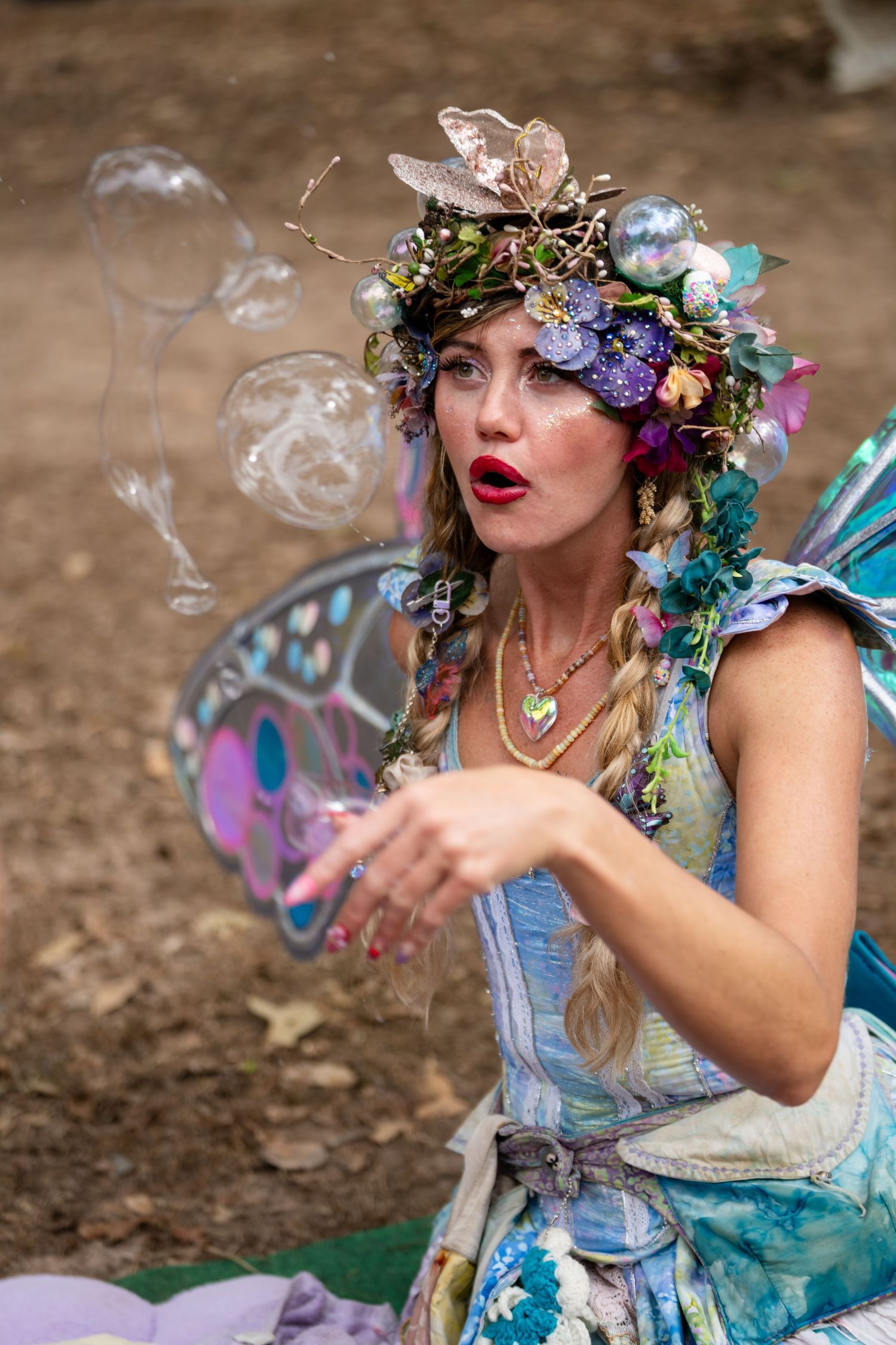 A whimsical fairy-like figure in blue dress and floral crown blows smoke rings while posing in a fantasy-themed photoshoot.