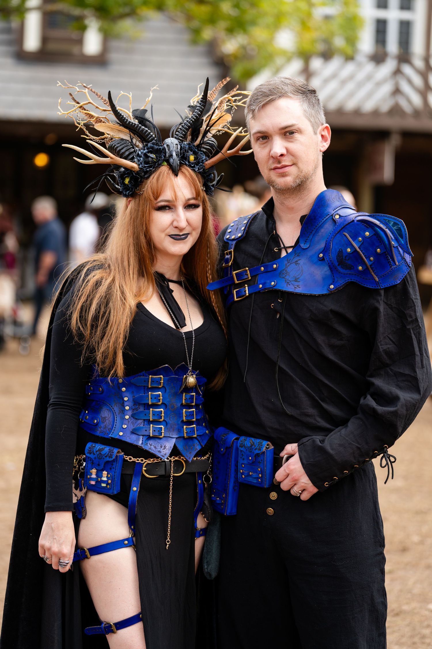 A couple in matching medieval fantasy costumes with blue leather corsets and black clothing at a renaissance faire.