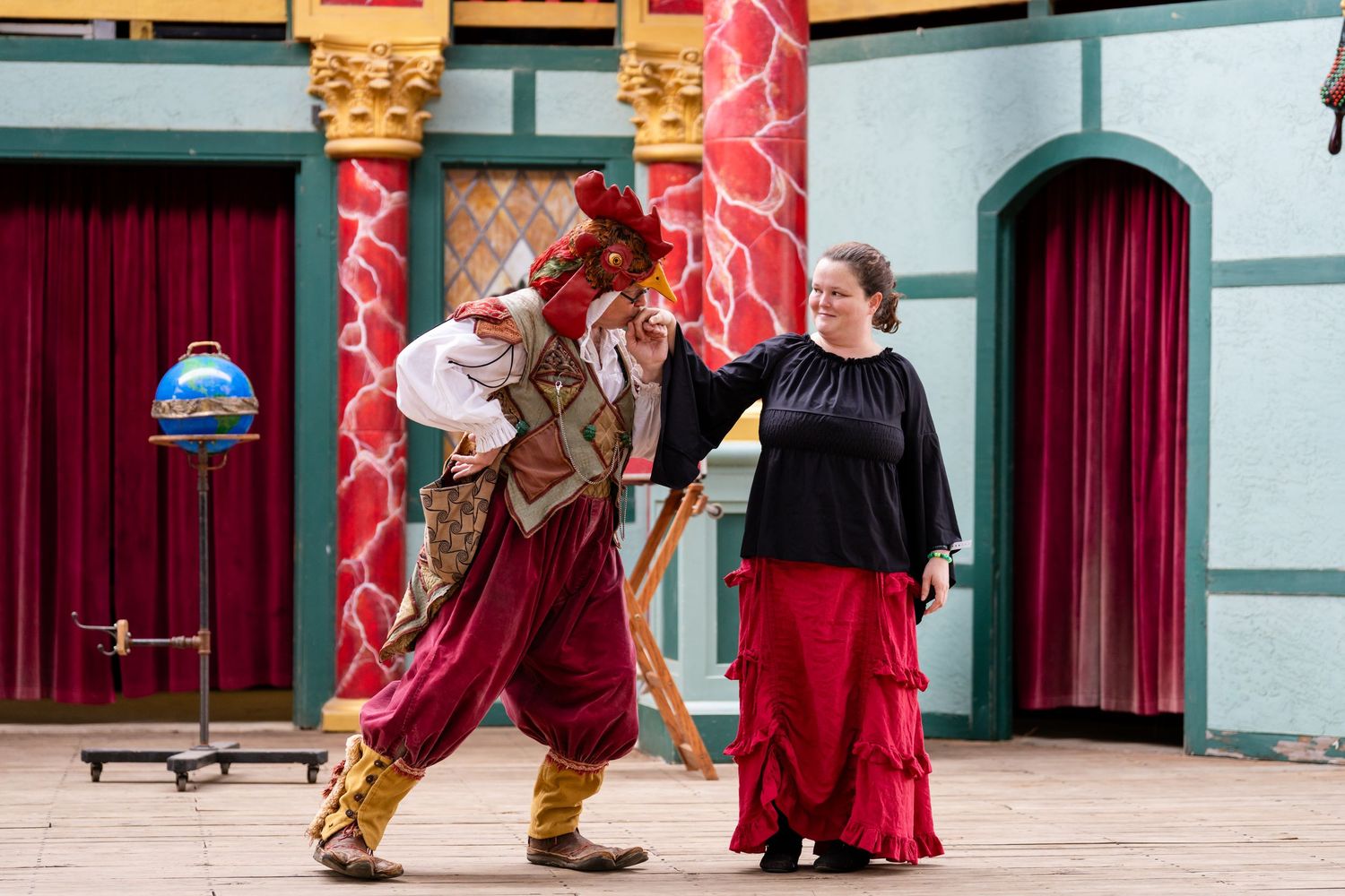 Two performers in period costumes act out a scene on a colorful stage with red curtains and decorative columns.