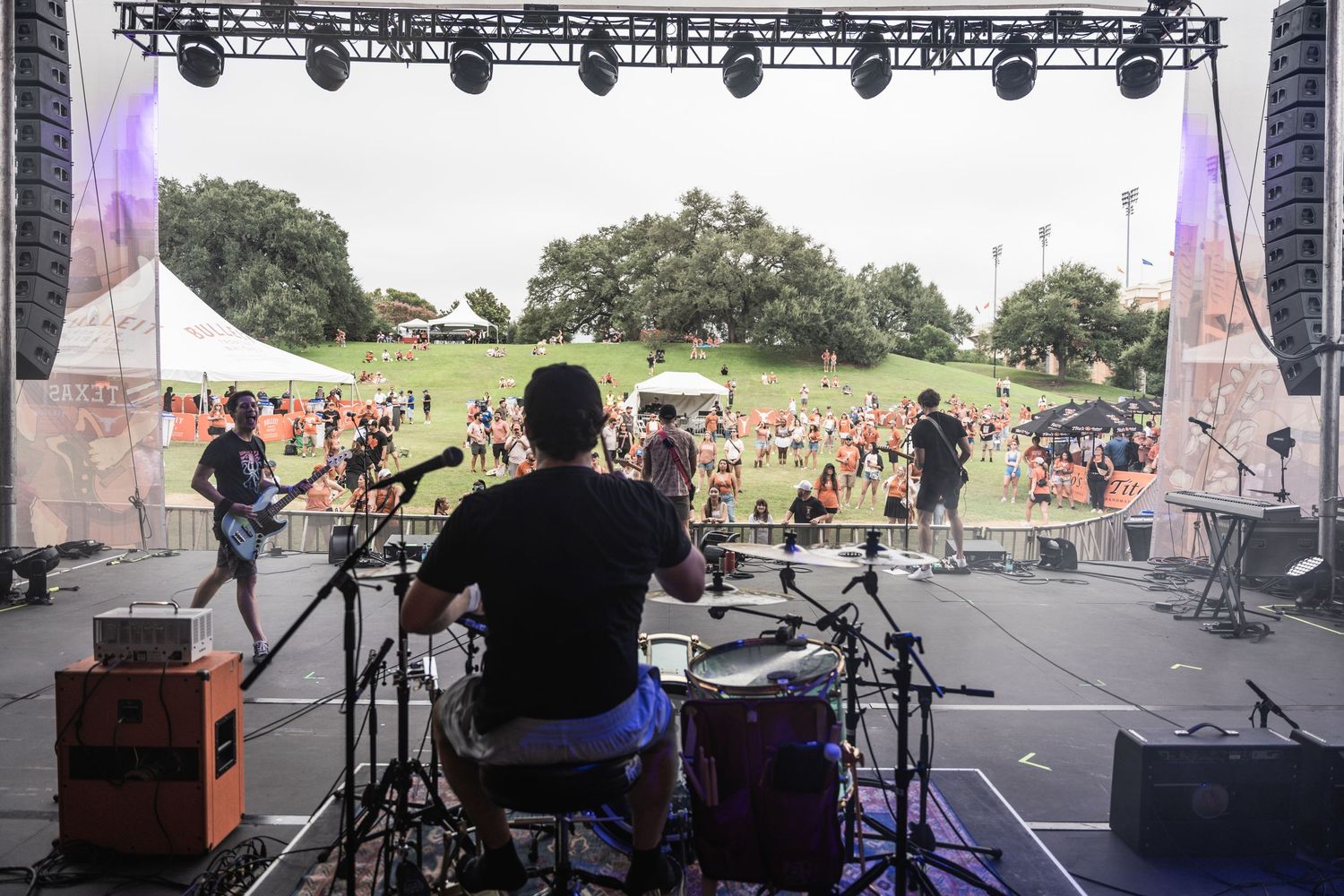 View from behind a stage setup showing lighting rigs and crowd at an outdoor music festival venue.