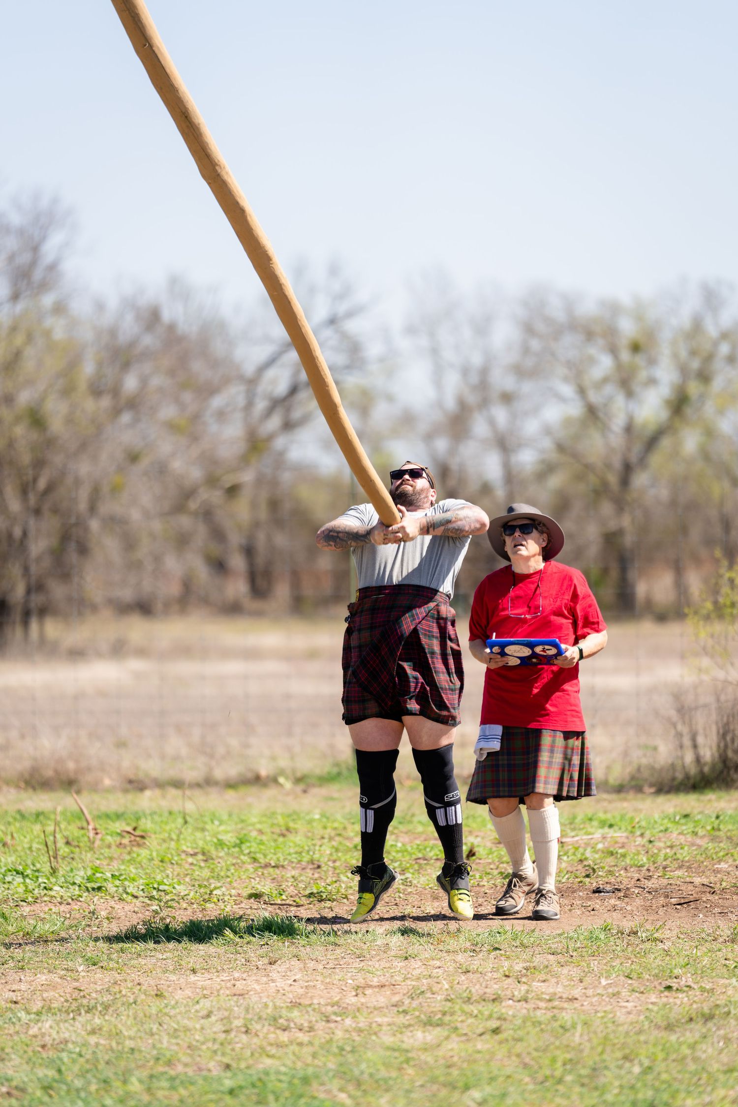 Man in kilt tossing a caber at a Scottish Highland Games competition while a judge observes nearby.