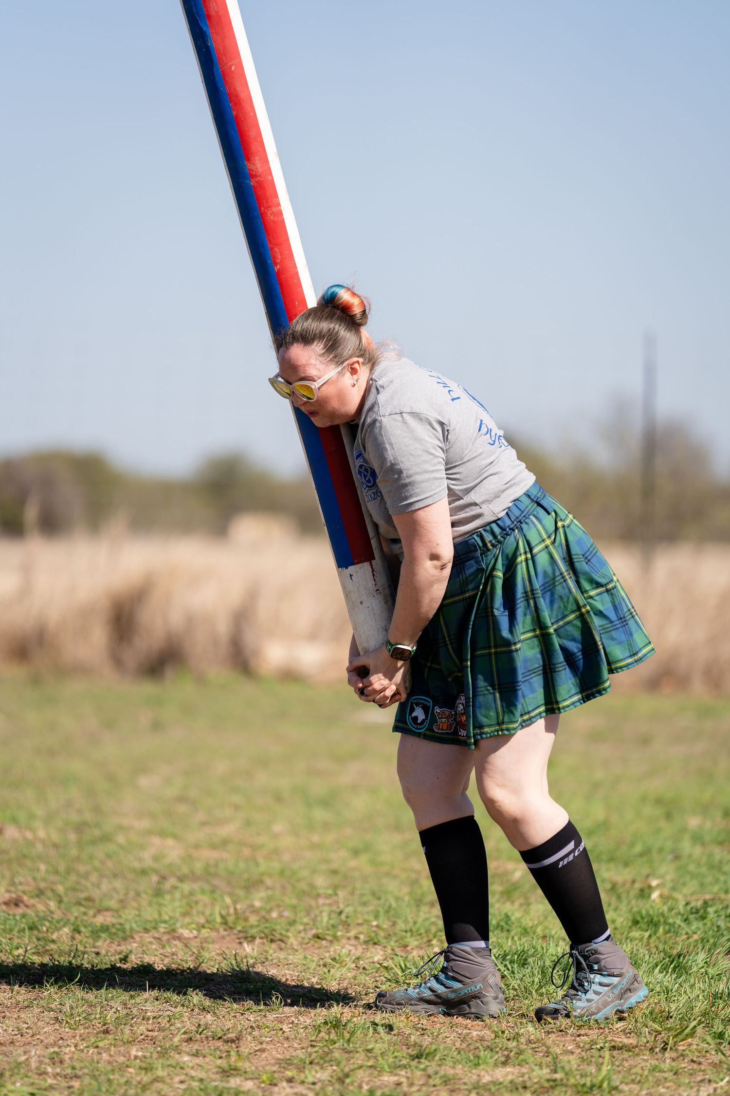 Woman in tartan skirt and yellow glasses tossing a caber at a Highland Games competition outdoors.
