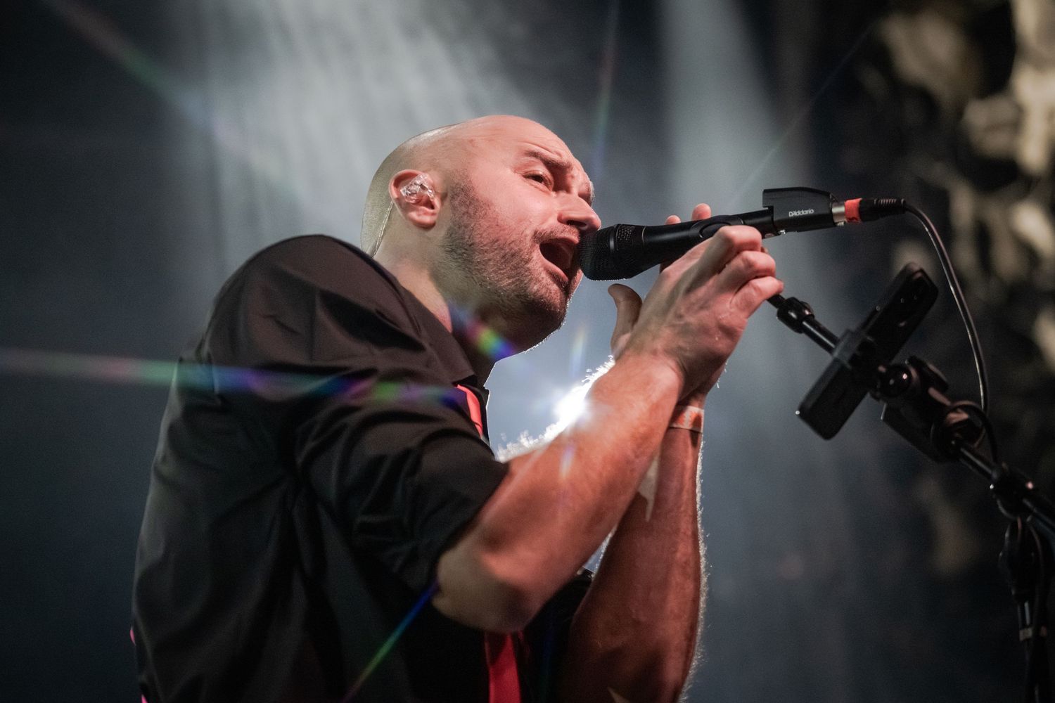 Bald male vocalist singing passionately into microphone on stage with dramatic lighting at live concert.