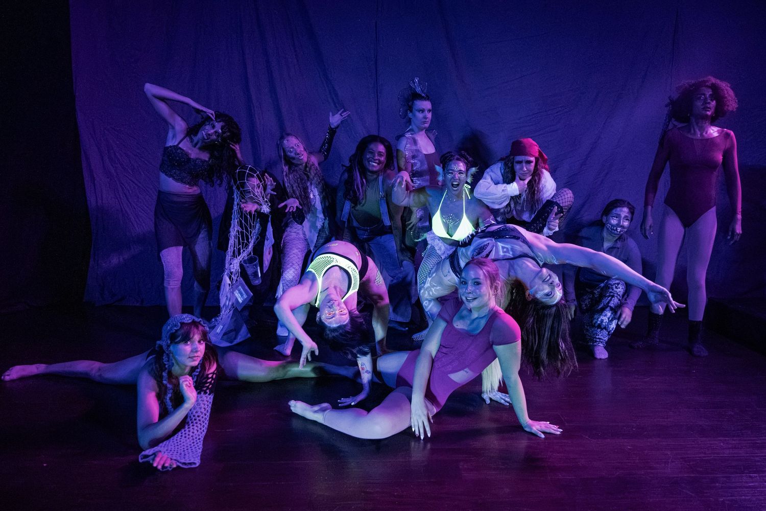 Diverse dance troupe performs dramatic group pose under purple stage lighting during theatrical performance.