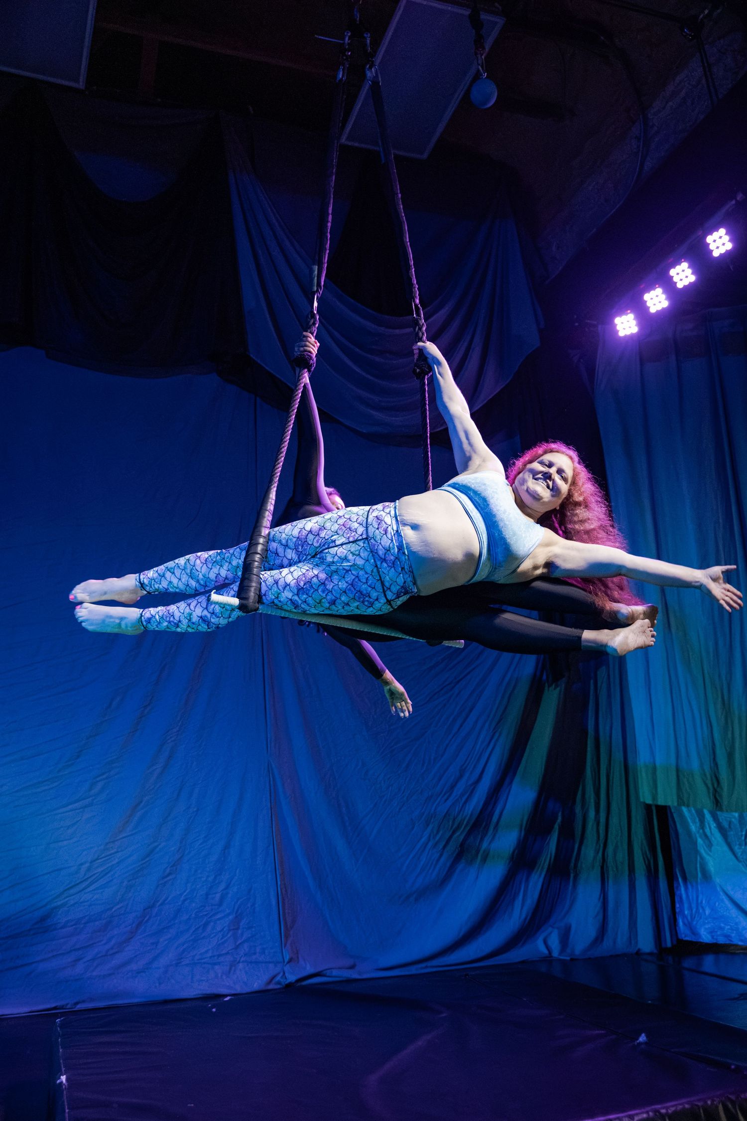 Aerial acrobat in mermaid costume performs graceful pose suspended on ropes against blue stage backdrop with purple lighting.