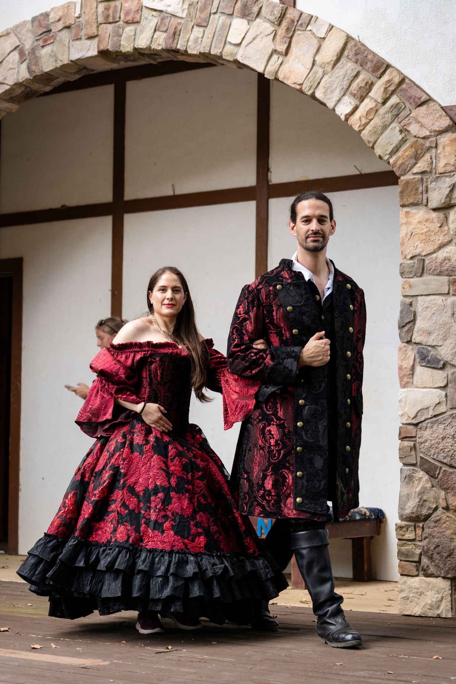 Couple in matching red and black Victorian costumes posing under a stone arch at a Renaissance faire.