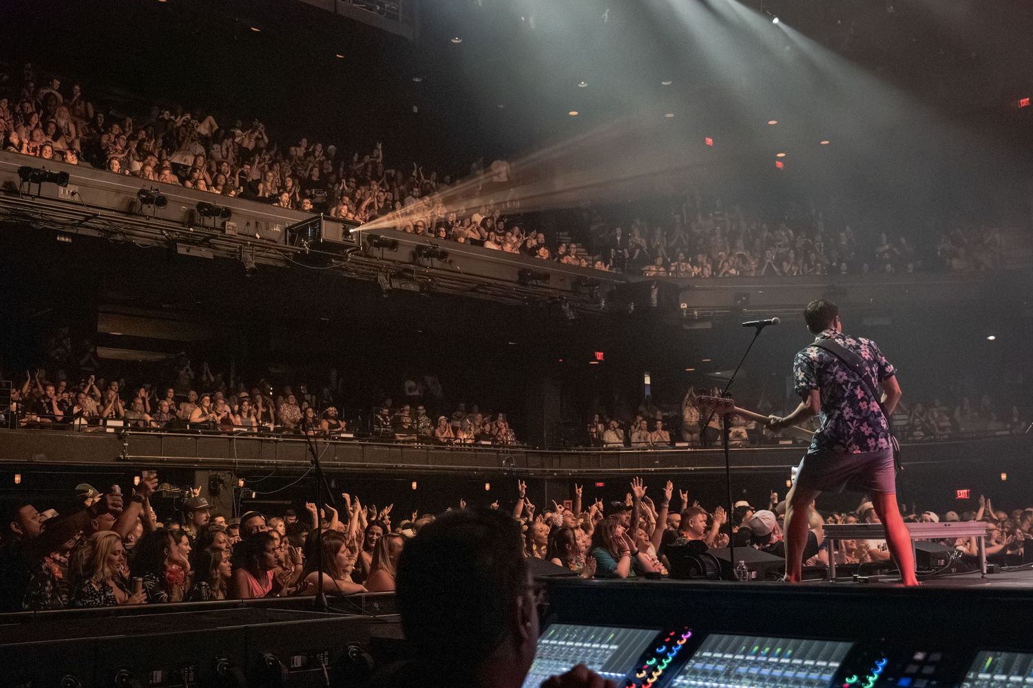 A rock performer on stage plays guitar for a packed concert hall crowd under dramatic spotlights.