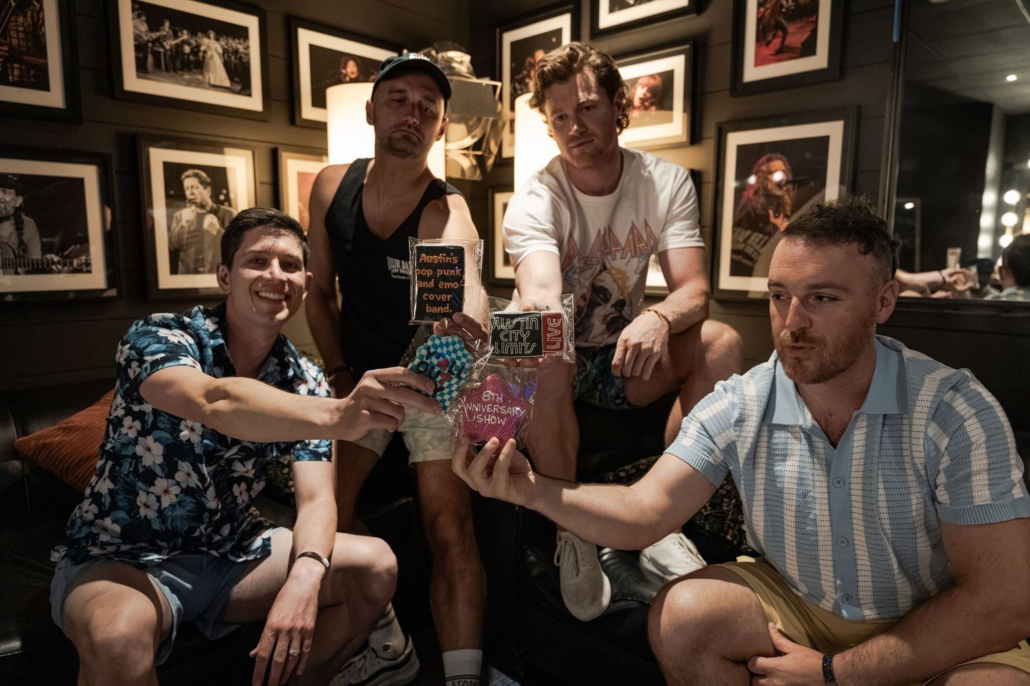 Four young men posing together in a room decorated with framed photos, holding drinks and smiling at the camera.