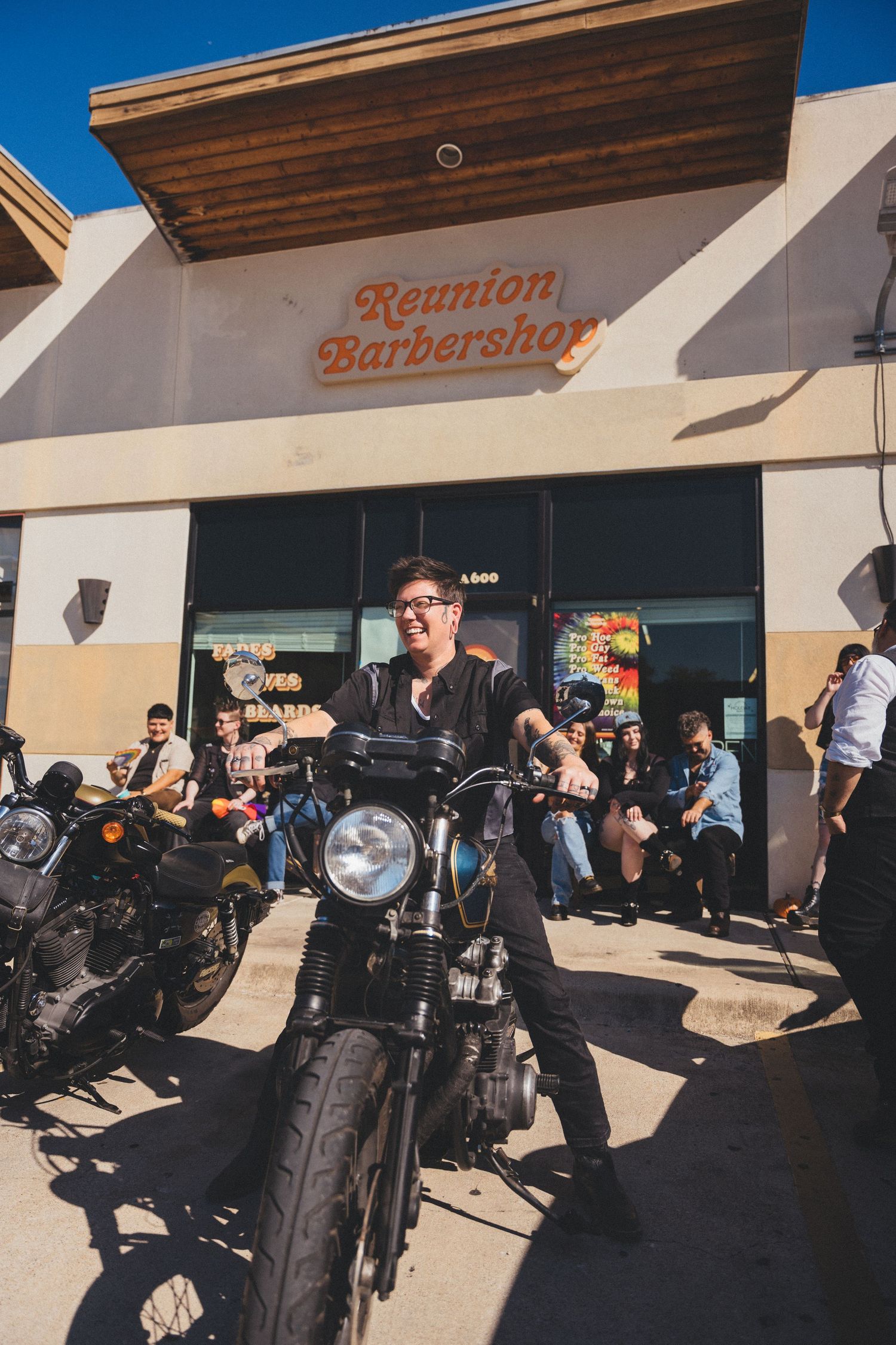 Motorcycle enthusiast poses on vintage bike outside Reunion Barbershop with crowd and other motorcycles in background.