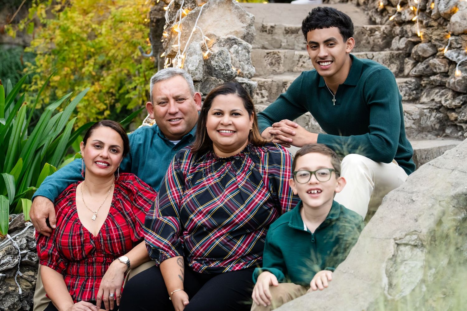Multi-generational family group sits together on stone steps surrounded by greenery during outdoor photo session.