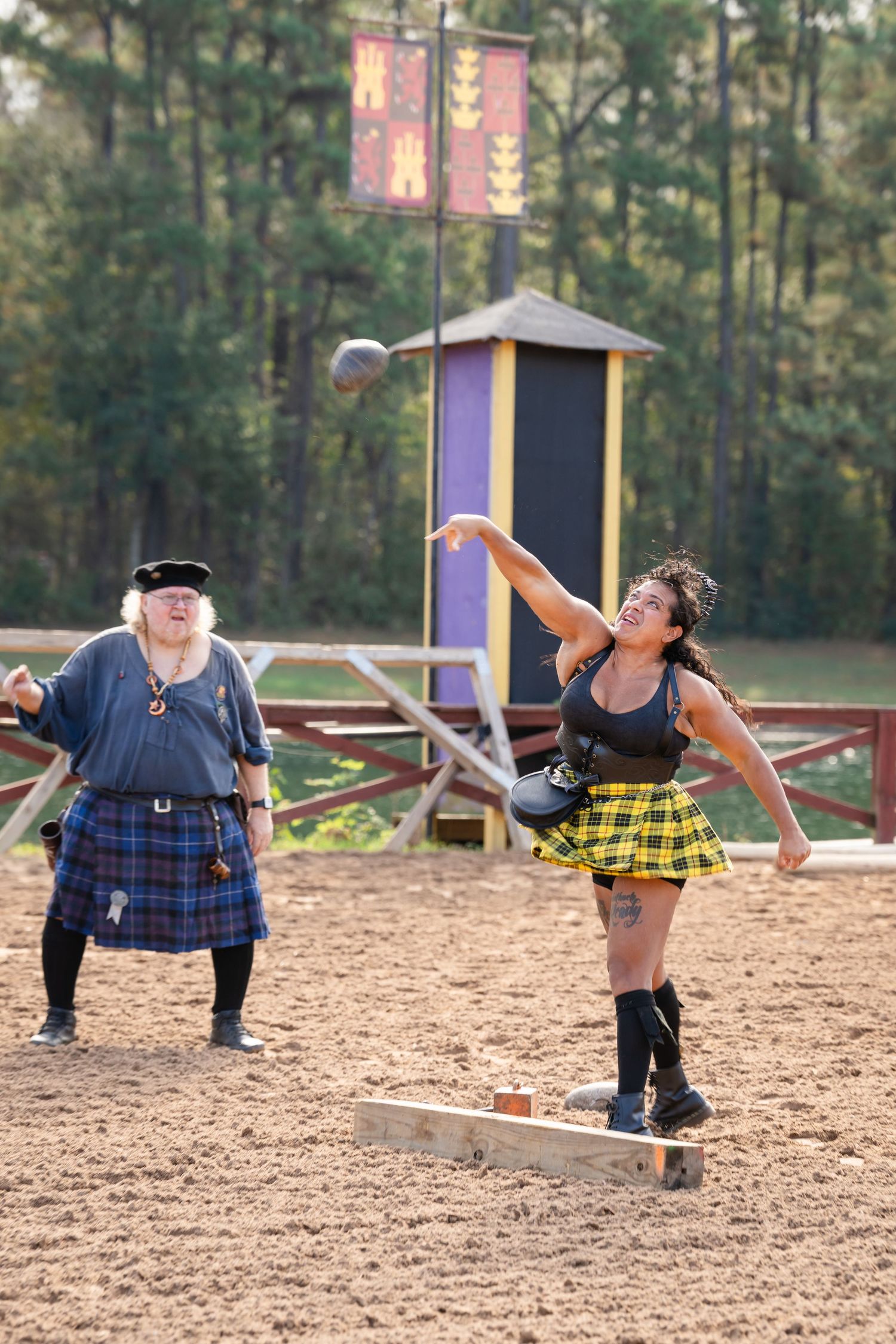 Athletes in kilts compete in a Highland Games throwing competition on a sandy field surrounded by pine trees.