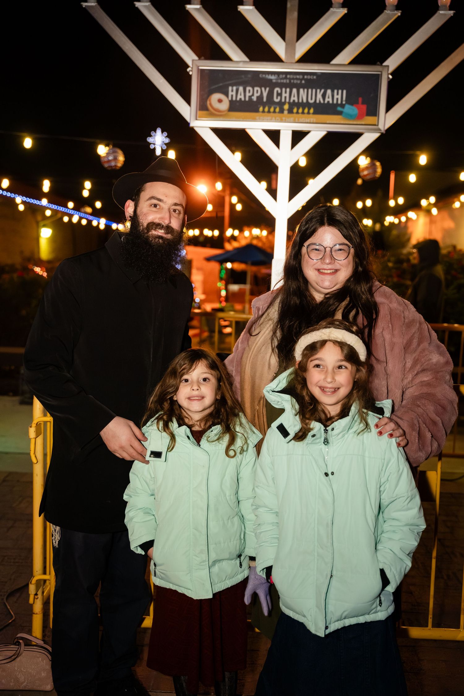 People gather at a Chanukah celebration with a large illuminated menorah and festive string lights in the evening.