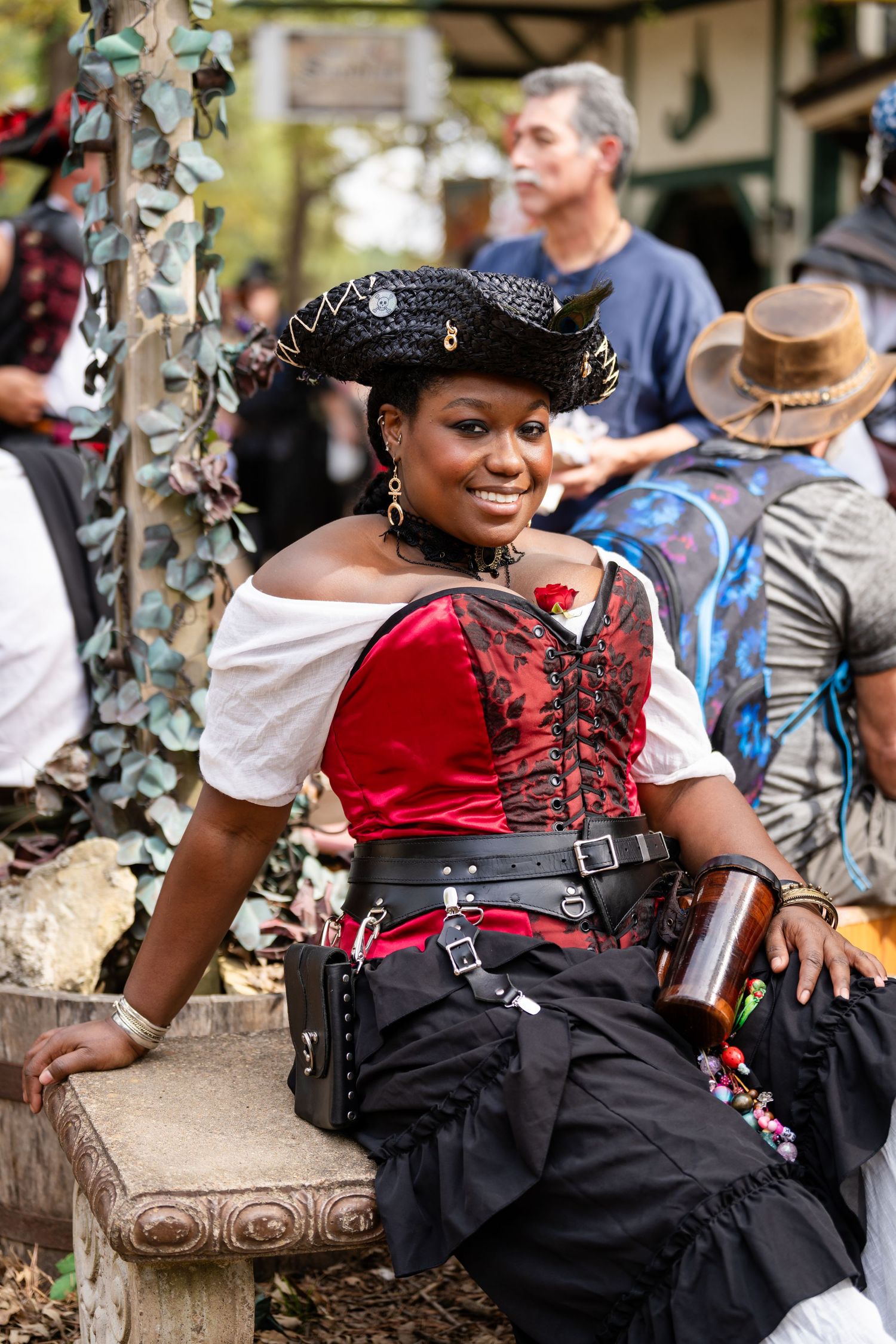 A person in renaissance festival attire wearing a black and red corset dress with decorative accessories sits outdoors.