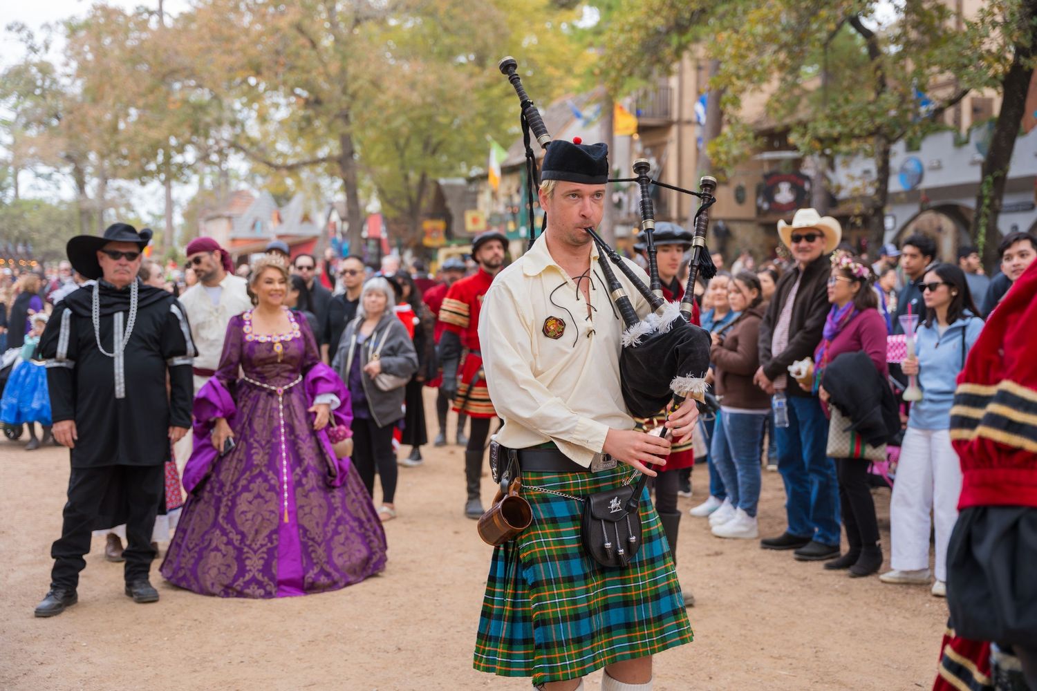 A bagpiper in traditional Scottish attire performs at a Renaissance festival while costumed attendees watch.
