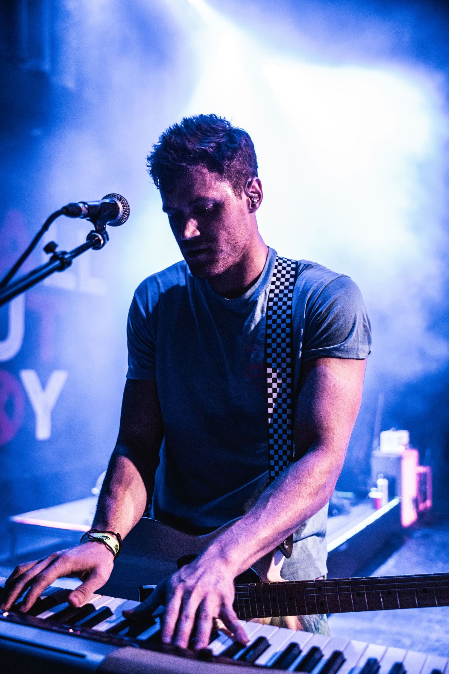 Musician plays keyboard wearing checkerboard strap under atmospheric blue stage lighting.