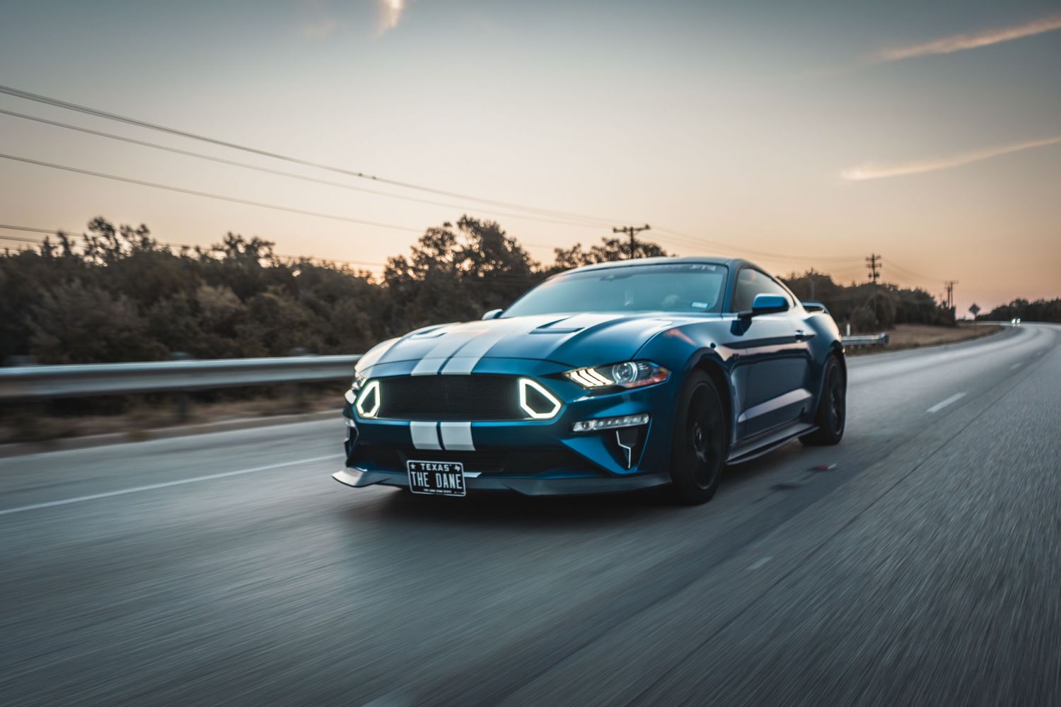 Modified blue Mustang with racing stripes photographed in motion during sunset.