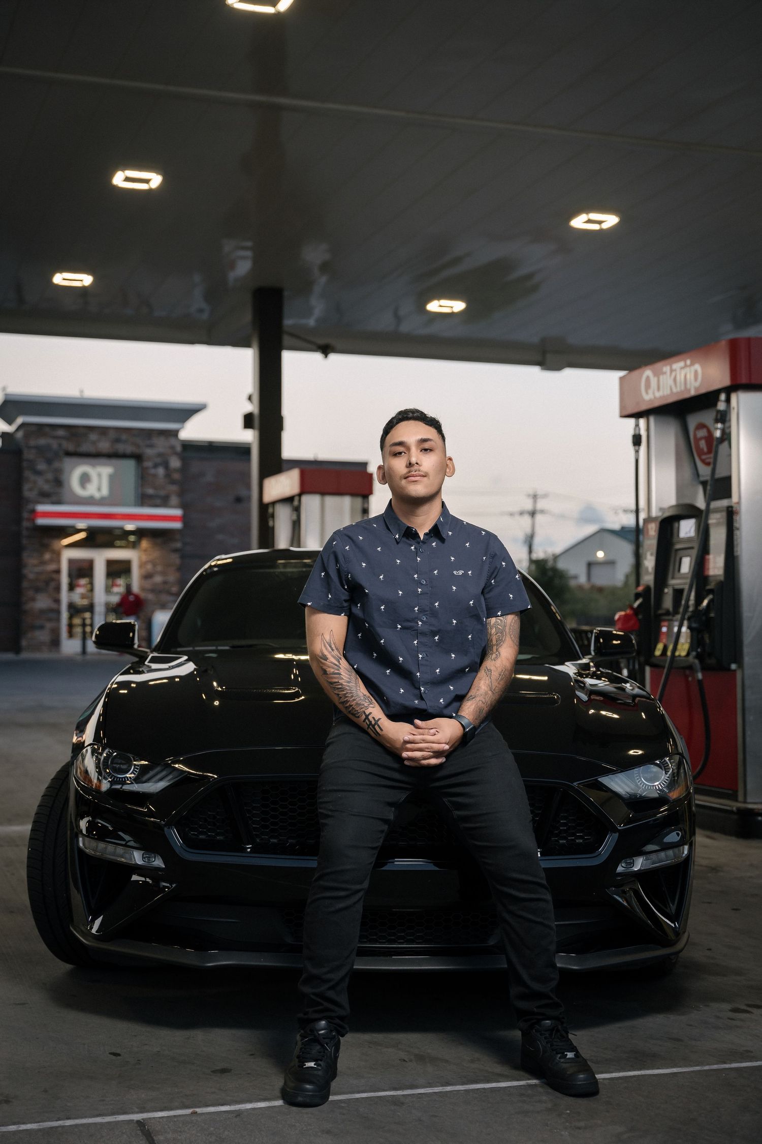 Stylish black Mustang and person in polka dot shirt at gas station at twilight.