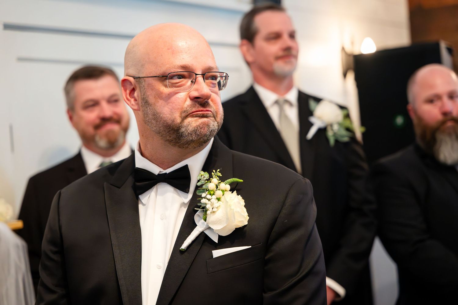Groomsman wearing black tuxedo with white boutonniere at wedding ceremony.