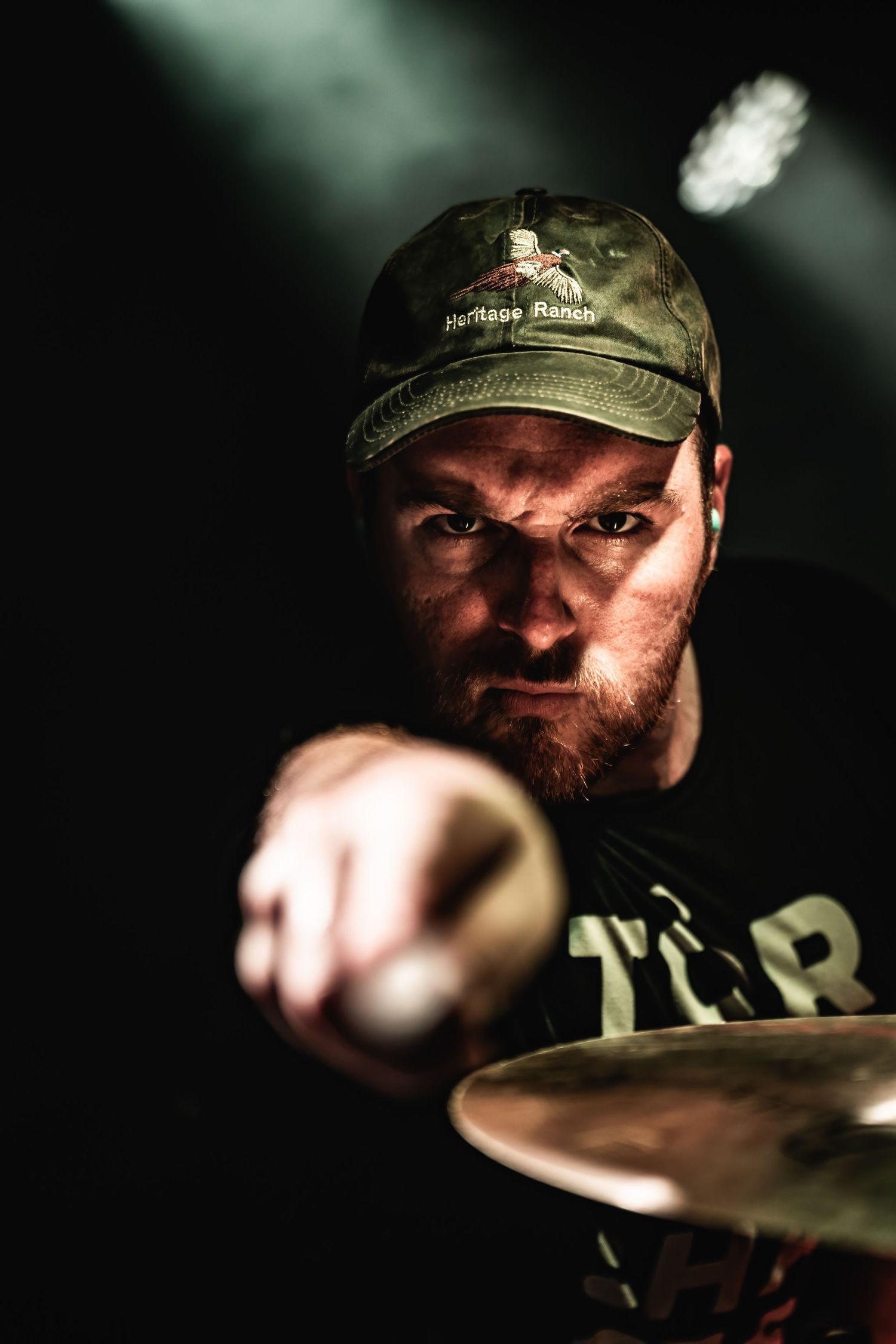 Dramatic portrait of a drummer performing in low light while wearing a camouflage cap.