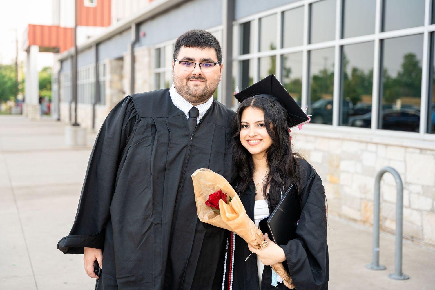 Two graduates in black caps and gowns pose together outside a building with glass windows.