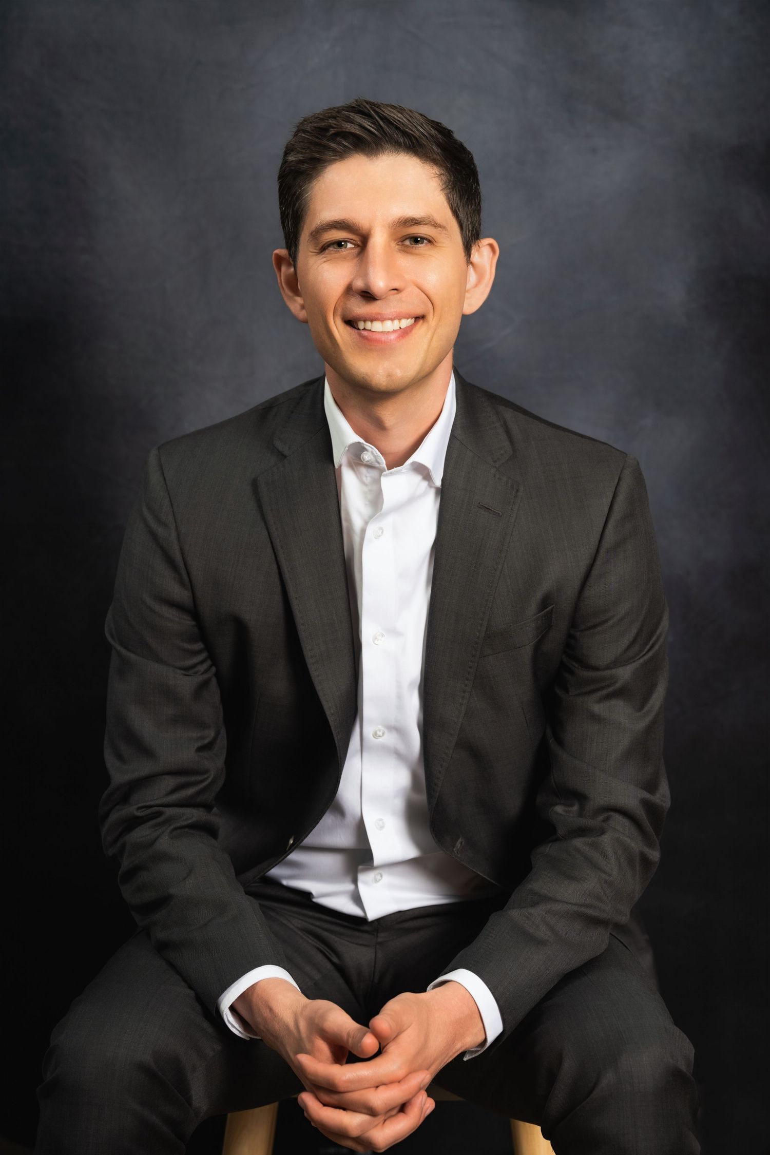 Professional business portrait of person in dark suit and white shirt seated against gray backdrop.