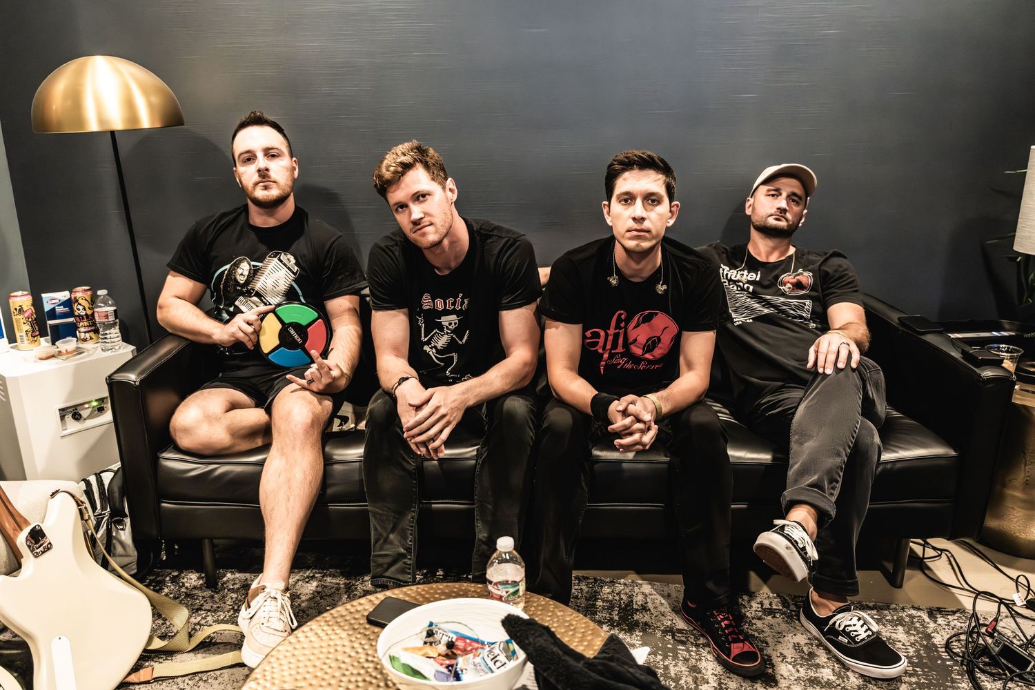 Band members sit casually on a black couch in a backstage lounge area.