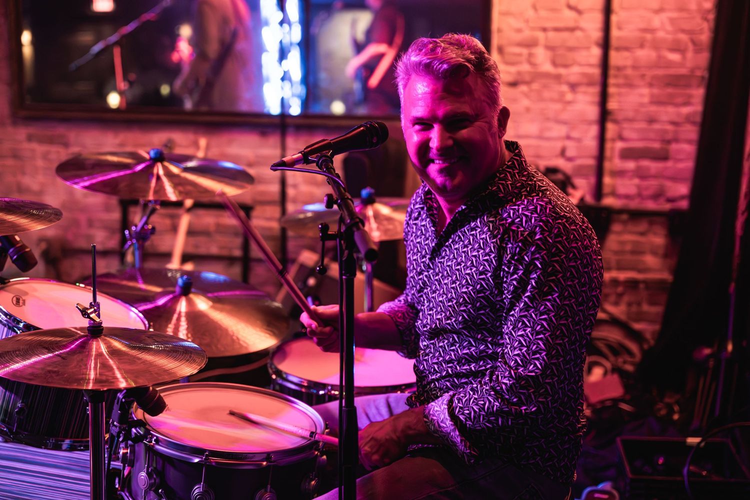 Drummer performing on stage with red and purple lighting in a brick-walled venue.