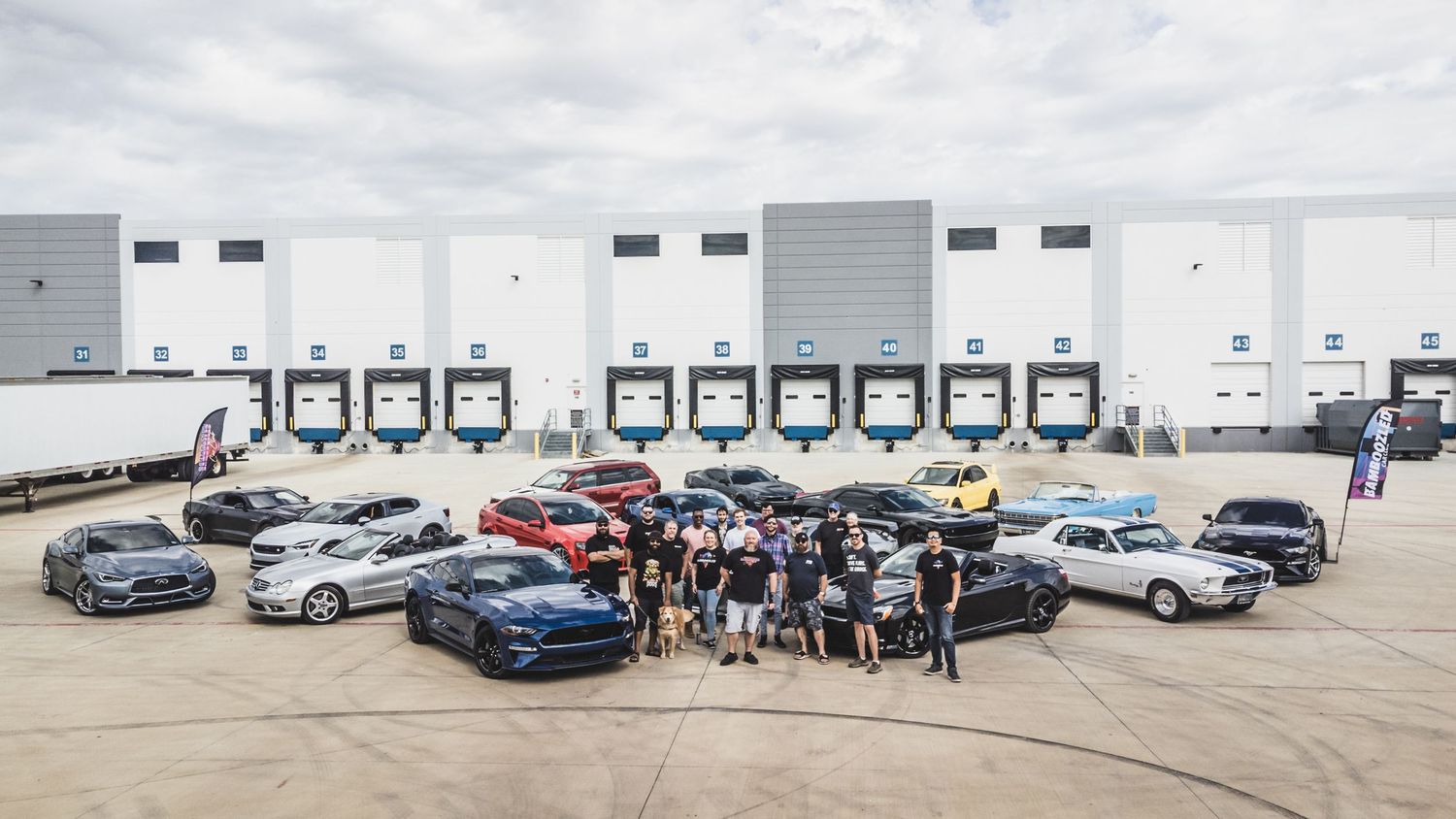 Large group of car enthusiasts and their vehicles gathered at an industrial warehouse location.