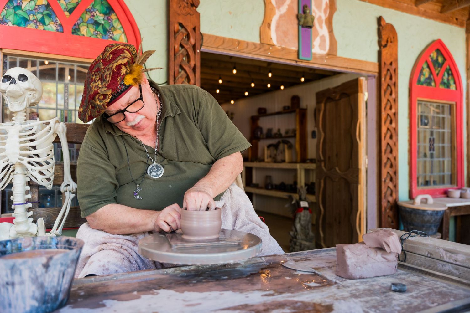 Artisan works on pottery at a medieval fair booth with decorative arched windows.