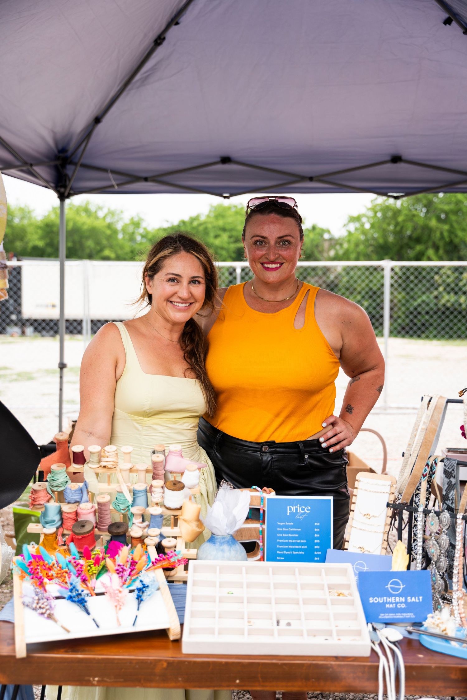 Two vendors stand behind a table displaying colorful candy, drinks and snacks under a white canopy tent at an outdoor event.