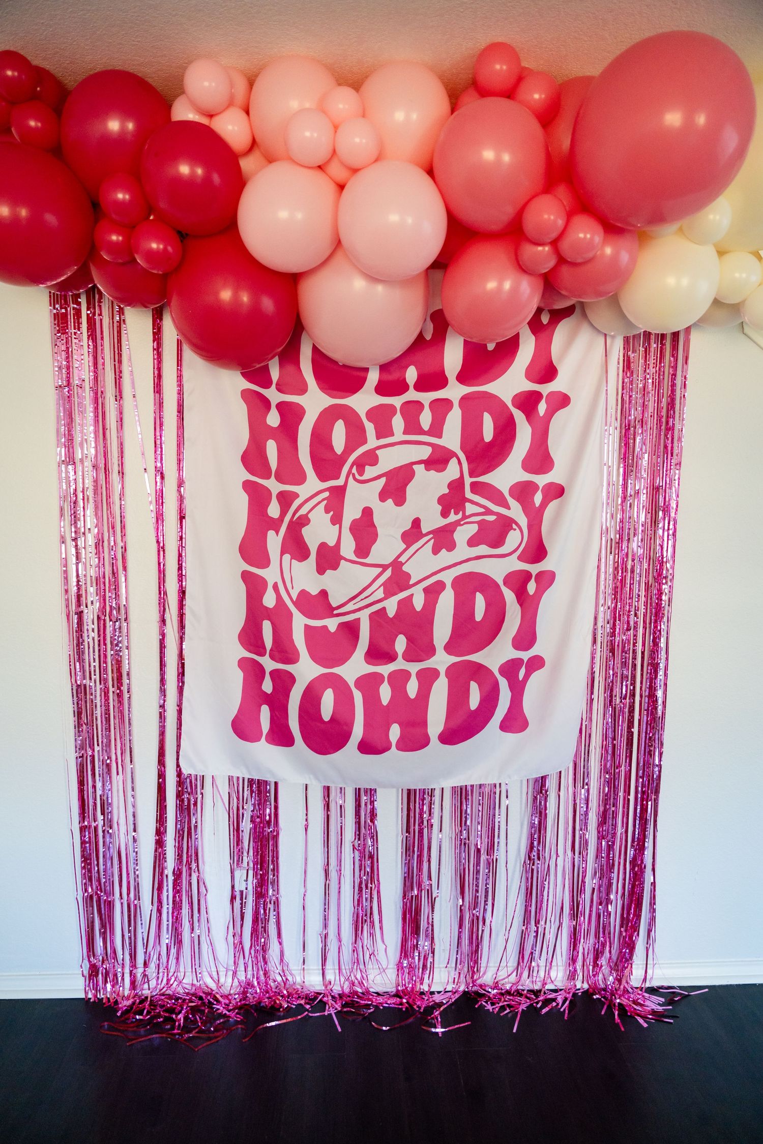 Pink party decoration setup with balloon arch and retro-style 'Howdy' text banner against metallic fringe backdrop.