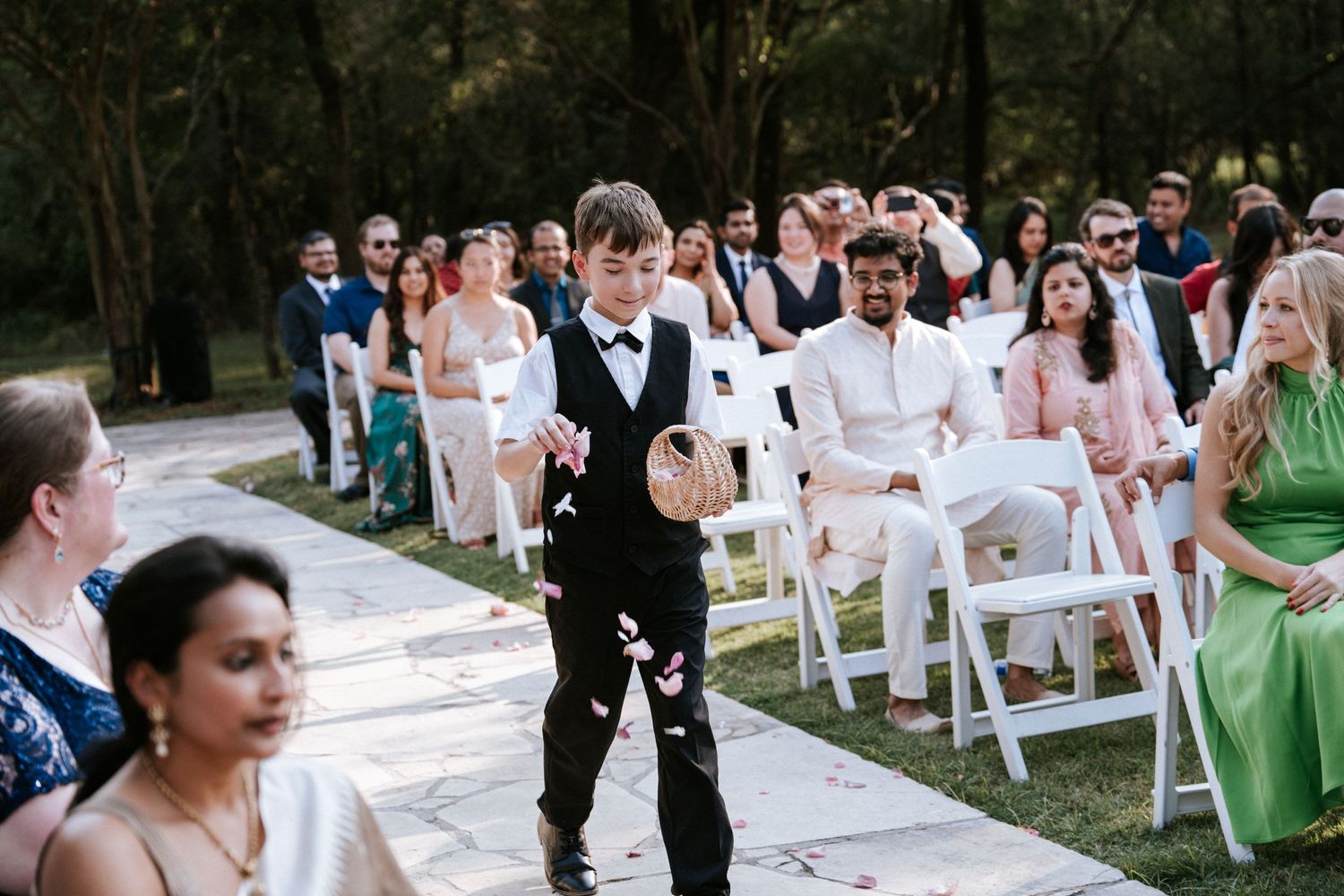 Guests seated in white chairs at an outdoor wedding ceremony with ring bearer walking down aisle.
