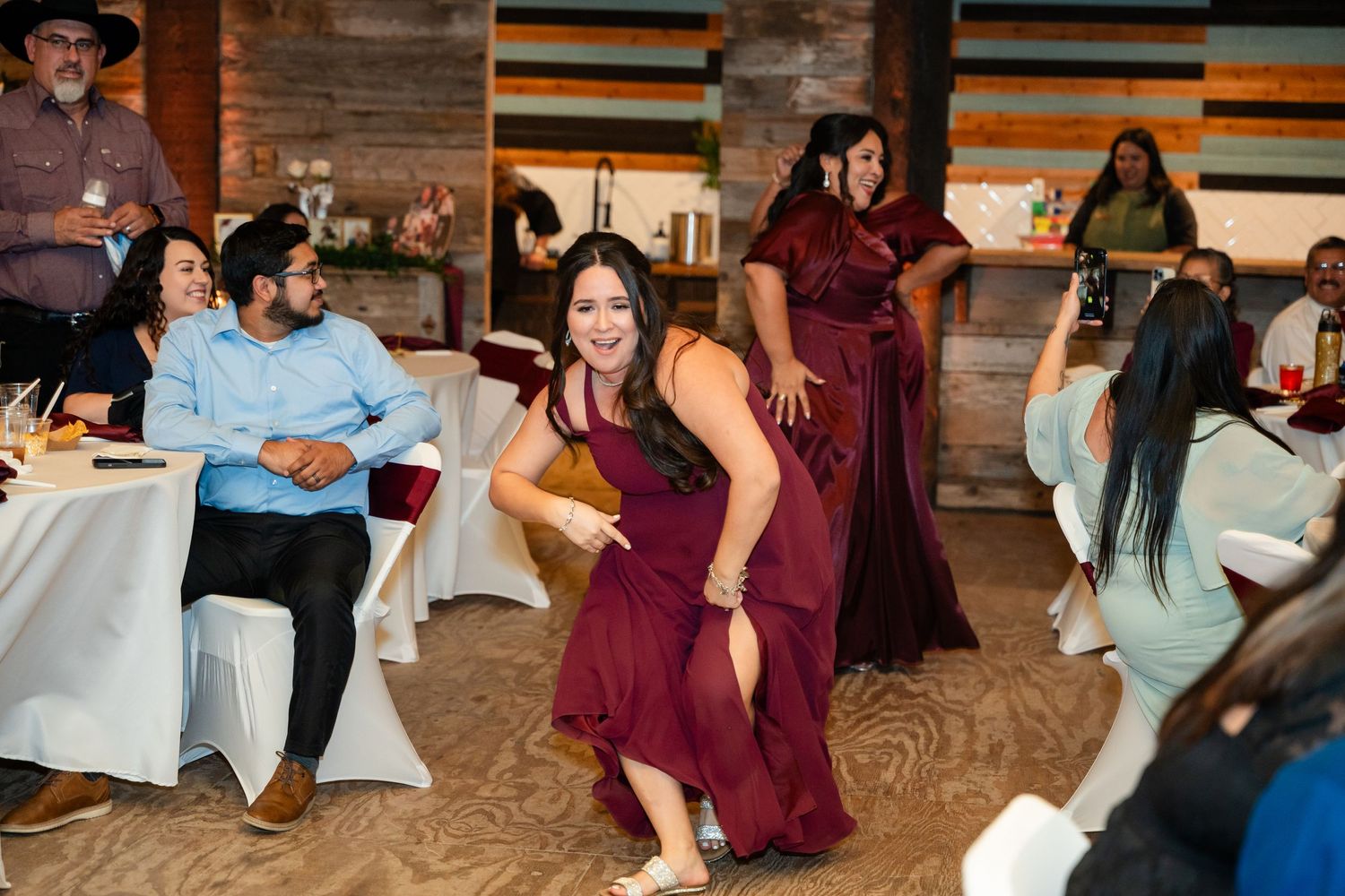 Wedding reception guests dancing and celebrating in burgundy attire at rustic venue.
