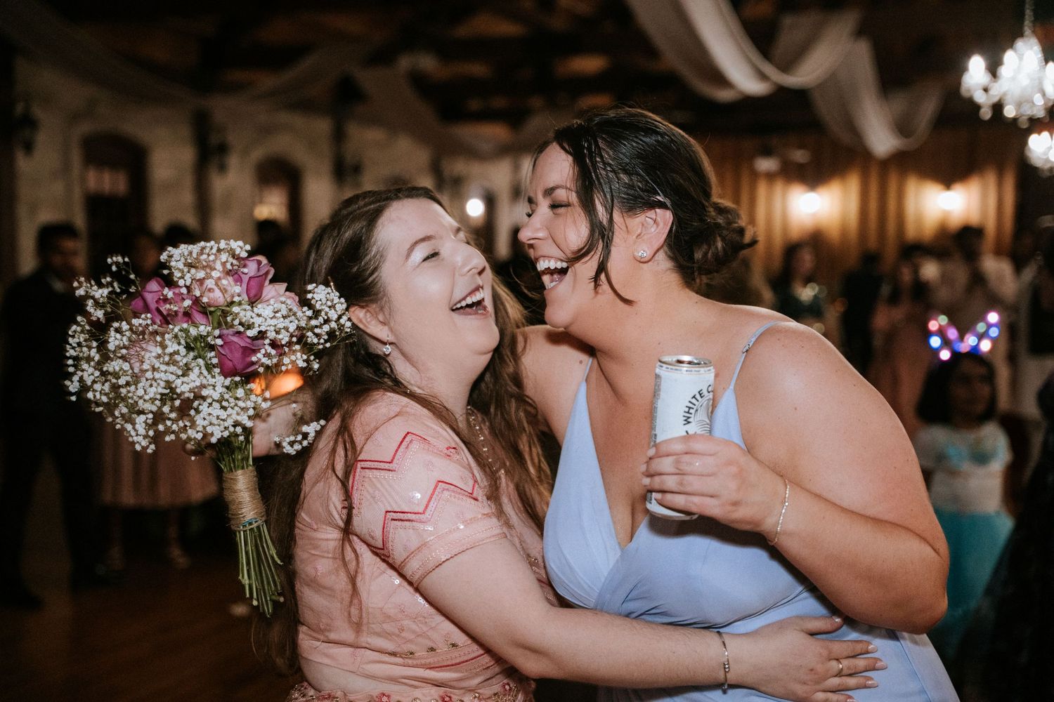 Two guests share a candid laughing moment while holding flowers at a wedding reception.