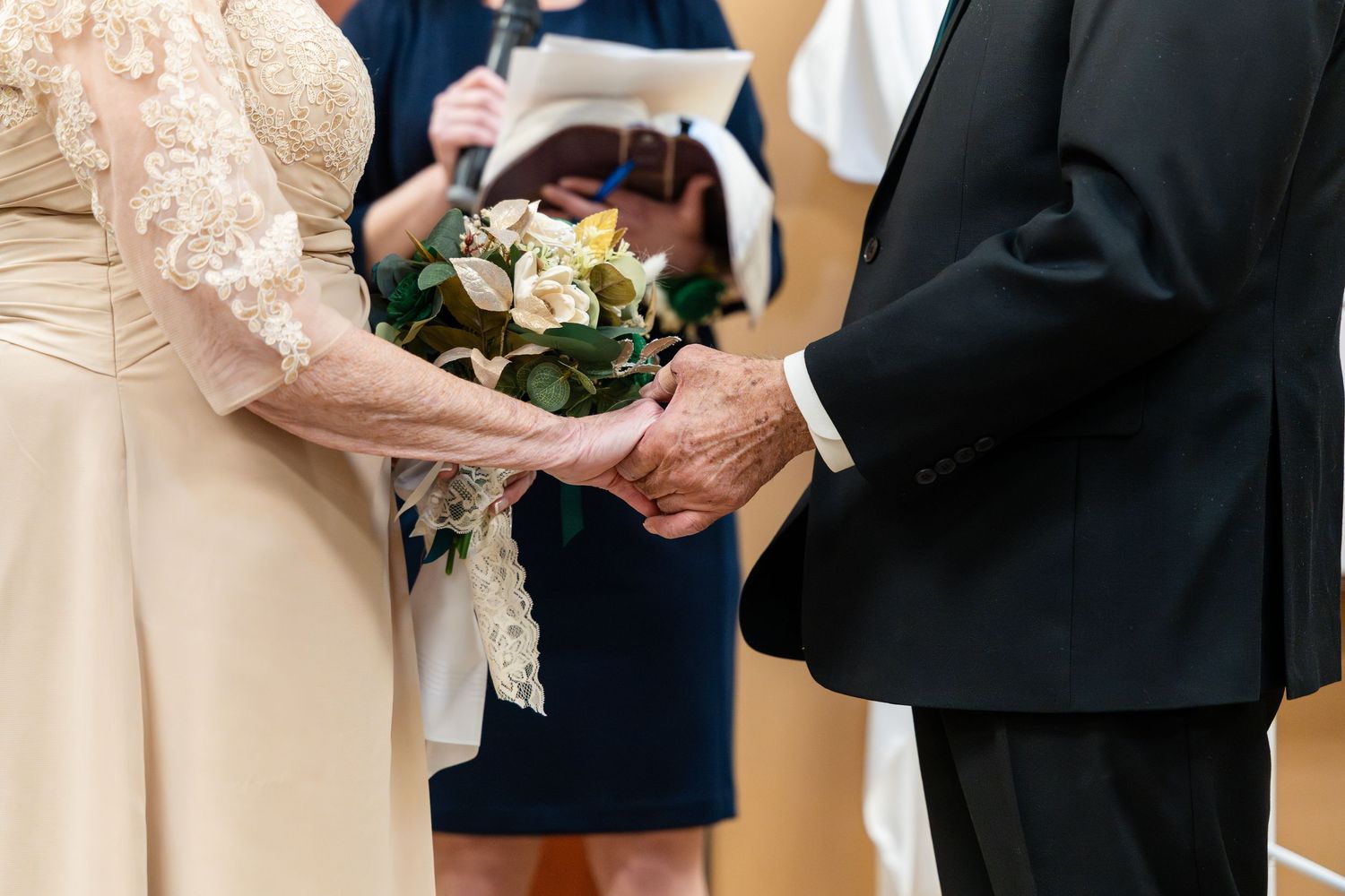 Close-up view of joined hands at a wedding ceremony with bouquet of white roses and cream-colored lace dress details.