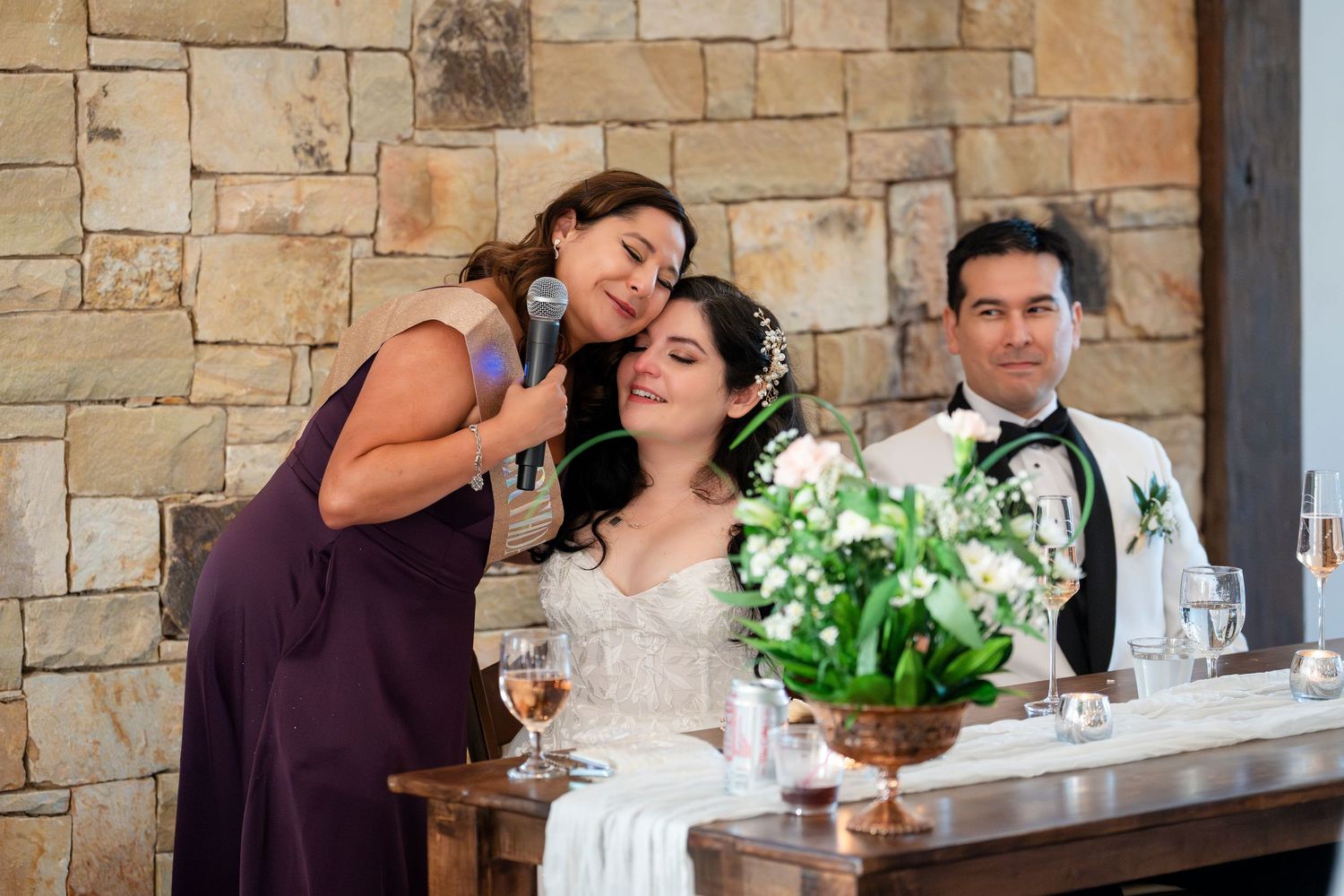Wedding party sits at reception table with stone wall backdrop and white floral centerpiece arrangement.